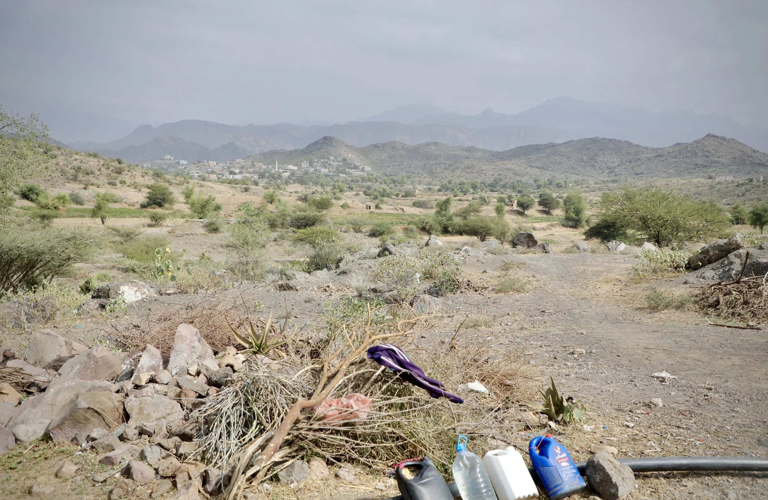 The view from the edge of Mosaid Mohammed Ali Naji's property, Shakhab, Al Dhale. The scrubland and hills visible beyond this boundary have not yet been fully cleared of landmines. Water containers and domestic objects mark the threshold between the 