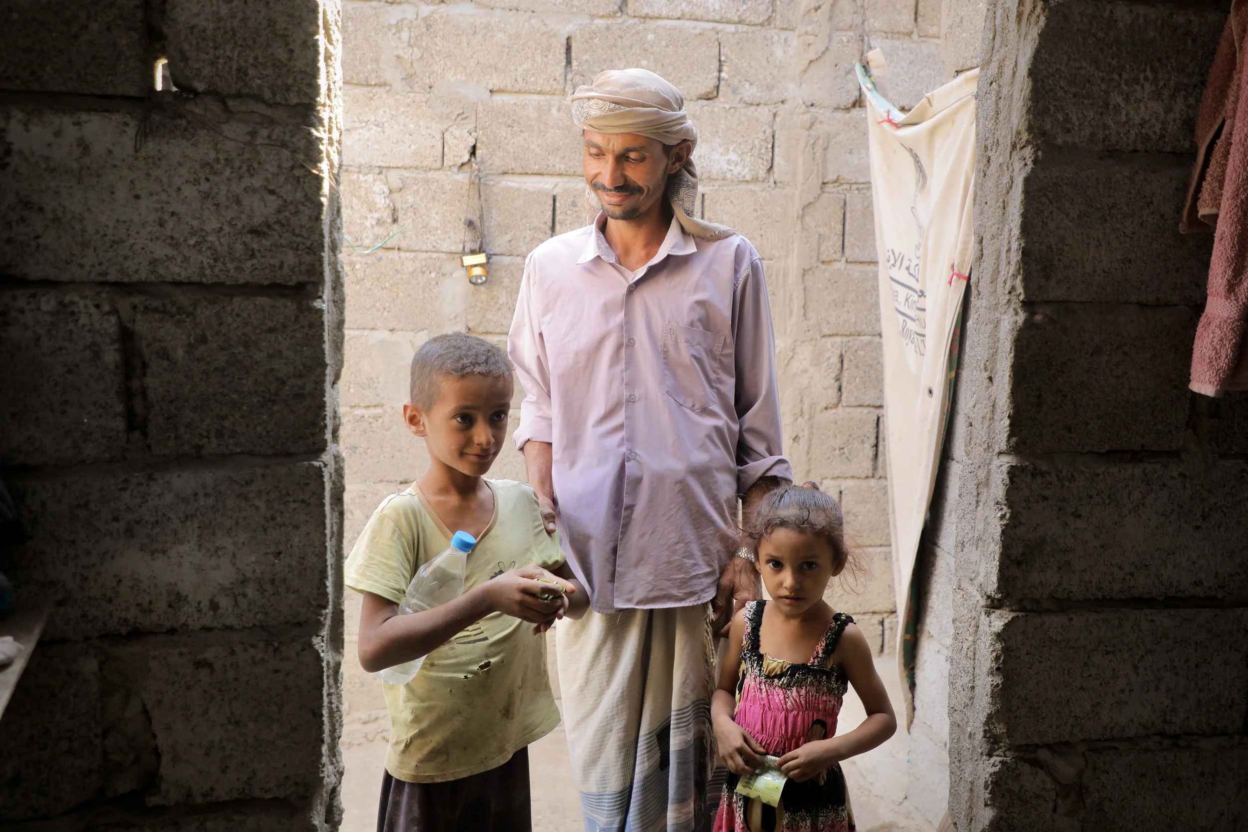 Amar stands with his children at the doorway of their home. Clearance has allowed the family to return and rebuild, though nearby land remains out of reach.