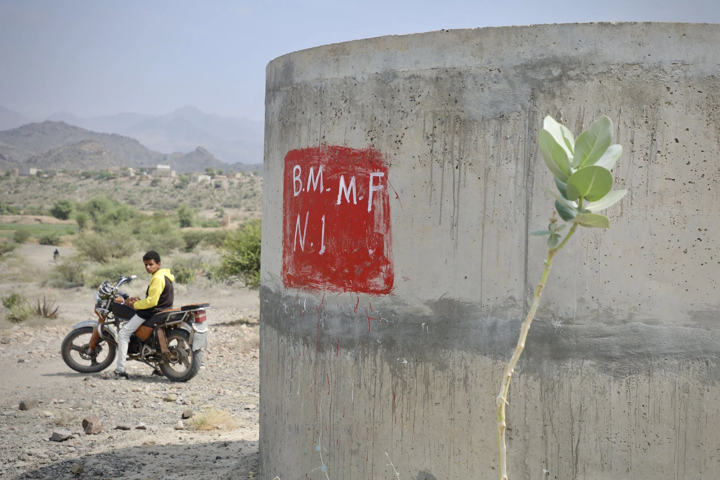 A Project Masam demining marker near Shakhab, Al Dhale. The red square indicates a cleared or active survey area. A boy passes on a motorbike. Life continues around the markers. The green sprig growing from the concrete is not incidental.