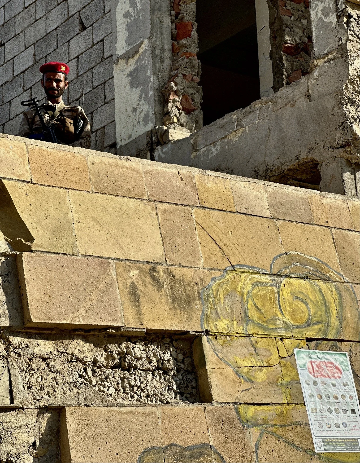 A soldier stands beside a damaged section of Ibrahim Aqeel School in Taiz, Yemen, where warning posters about landmines and improvised explosive devices are displayed on the walls.