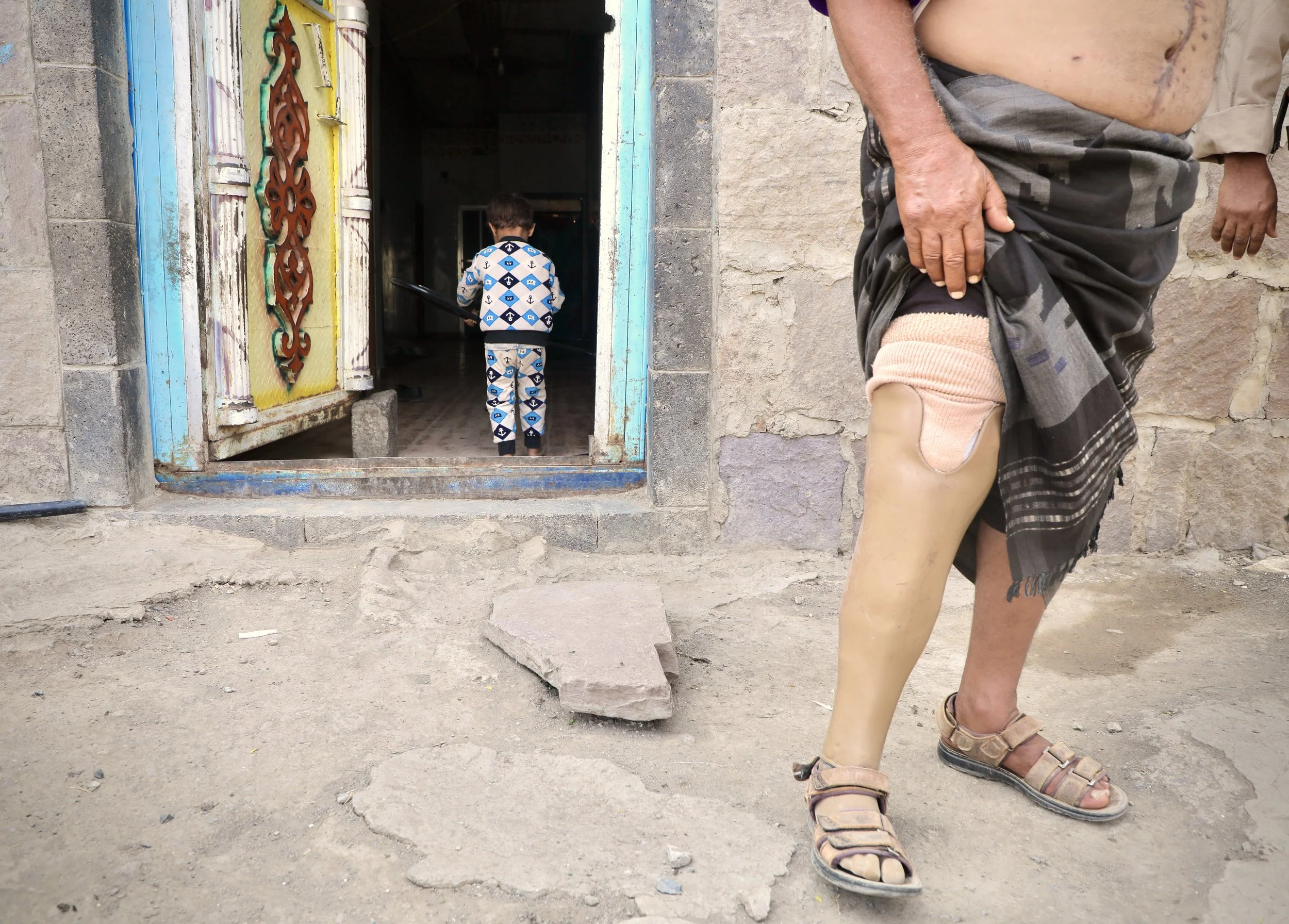Mosaid Mohammed Ali Naji, Shakhab, Al Dhale. His grandchild - wearing anchor-print pyjamas - walks toward the interior of the home. Mosaid stands on the ground of his courtyard, where a further 17 landmines were subsequently discovered. The burn scar