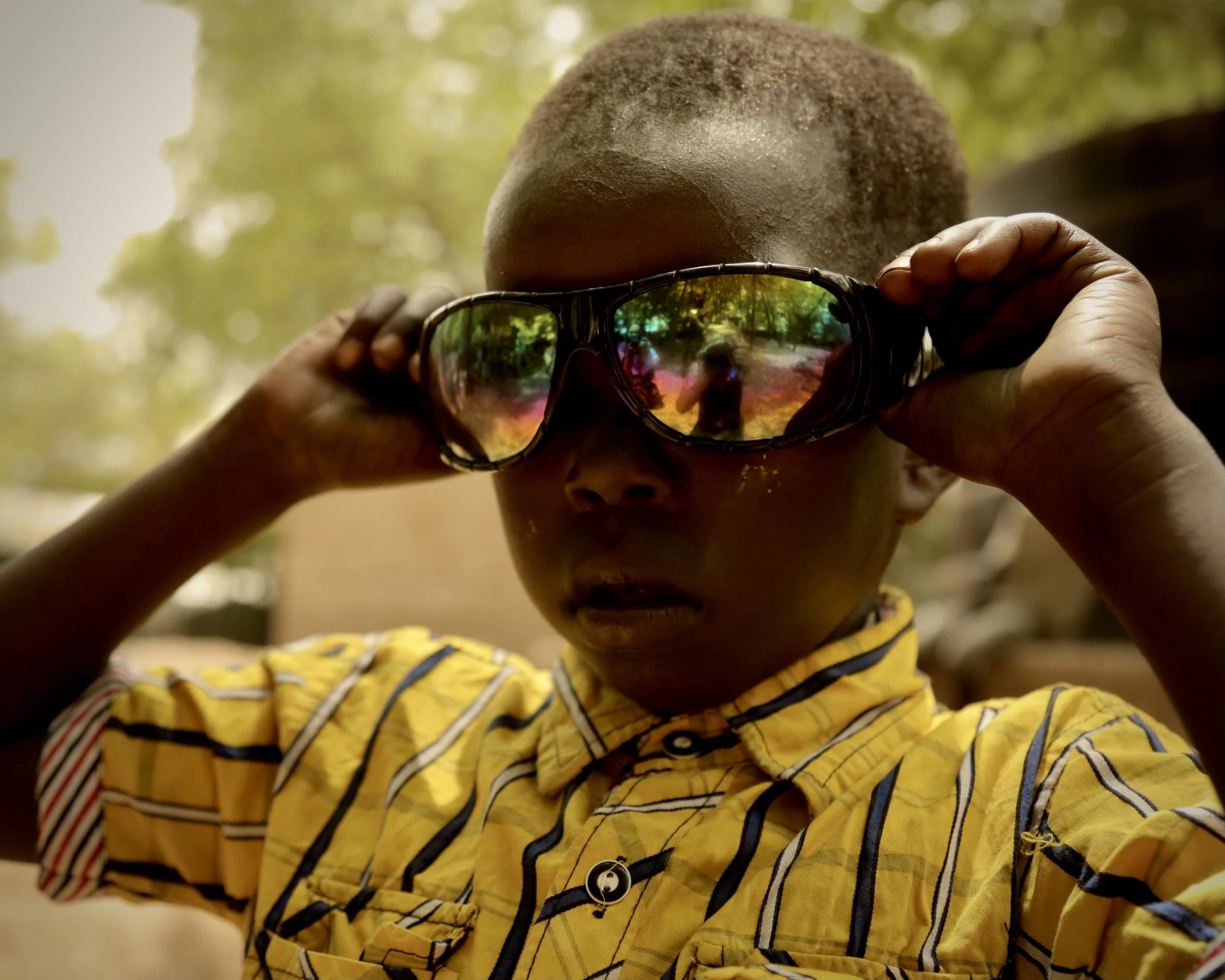 A South Sudanese boy in Kakuma refugee camp adjusts mirrored sunglasses. Faint traditional forehead scarification marks, practiced historically by several Nilotic communities, hint at family heritage carried across displacement.