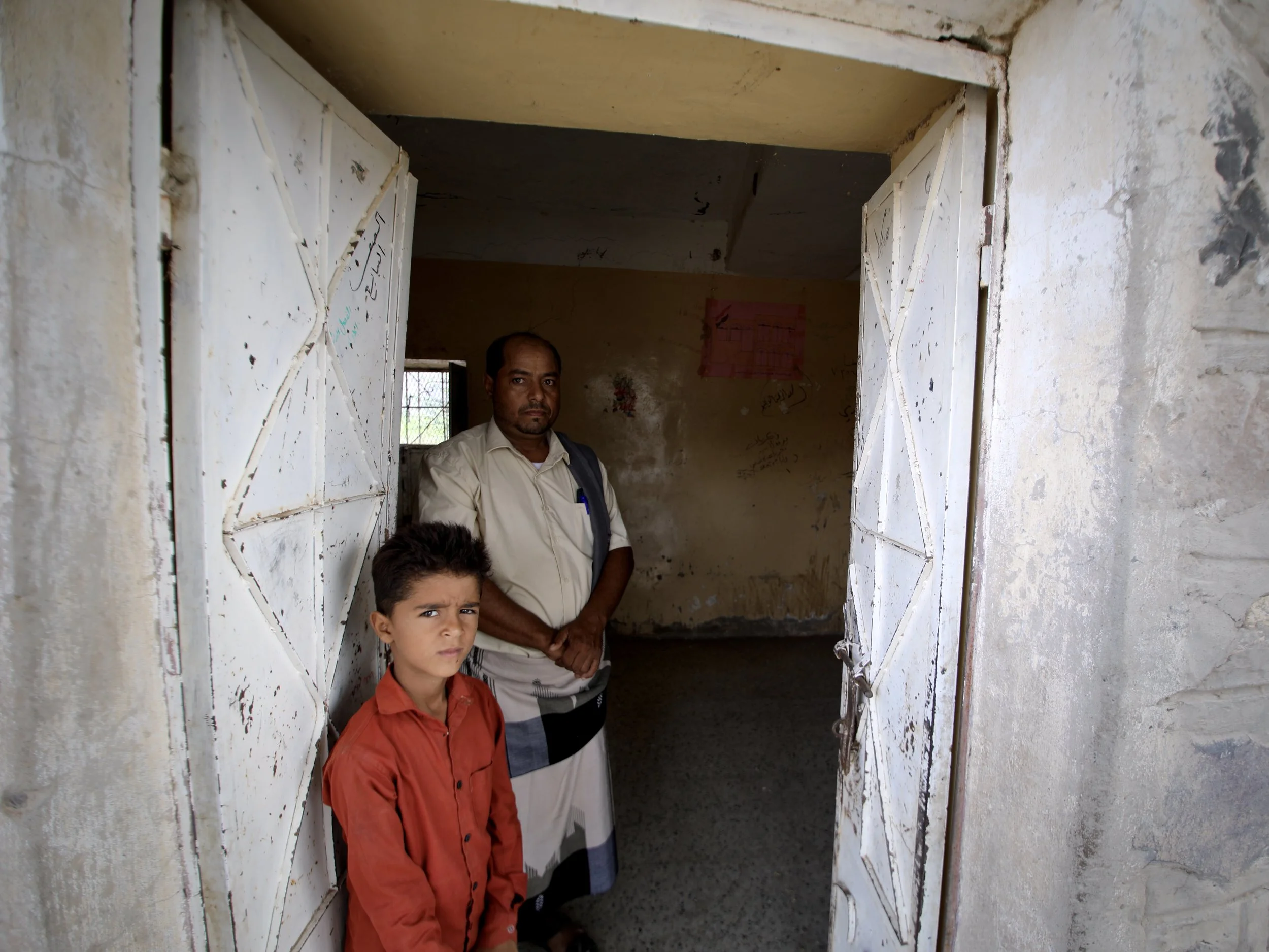 School Director Ahmad Mohamad Al Barakani, 33, and his son Marwan, stand in the doorway of a damaged classroom at Al Shaab School in Jabal Habashi district, Taiz governorate.