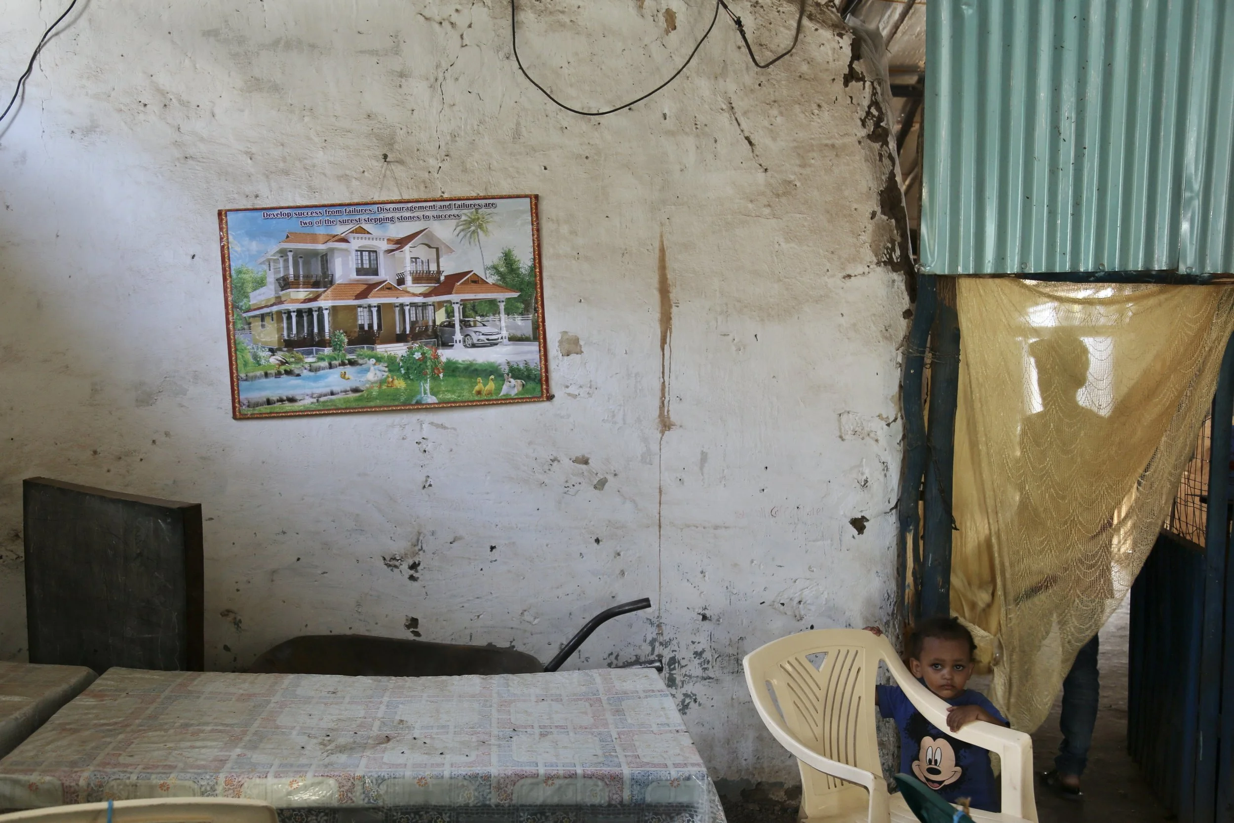 Nazrit’s younger brother sits beneath a poster of a large house fixed to the wall. Behind the curtain, Nazrit stands in shadow: an image of hope, waiting and the future imagined from inside the camp.