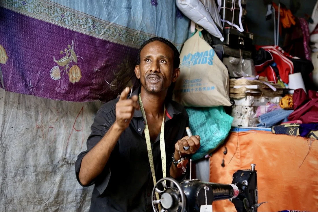 The tailor gestures while speaking with visitors who spend time in the shop.