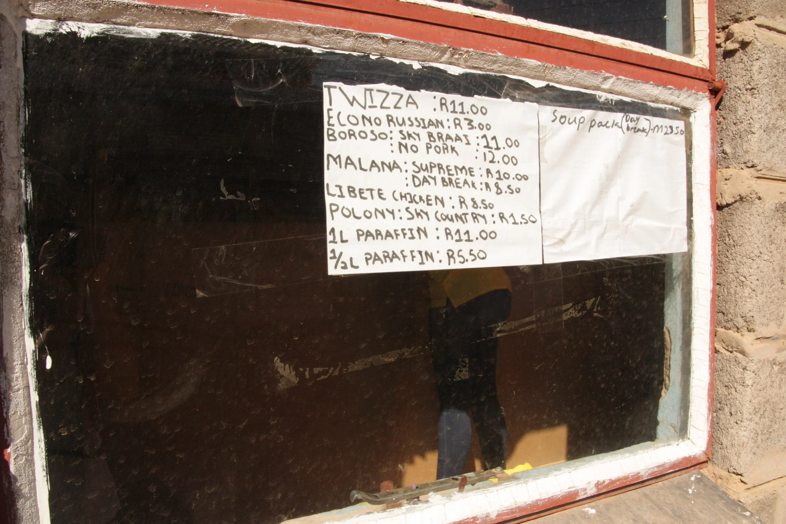 Seen through the dusty shop window, a woman waits at the till behind handwritten price lists outside Paballo’s store in Lesotho. The photographs avoid fully identifying Paballo in order to protect her privacy and safety while documenting the environm