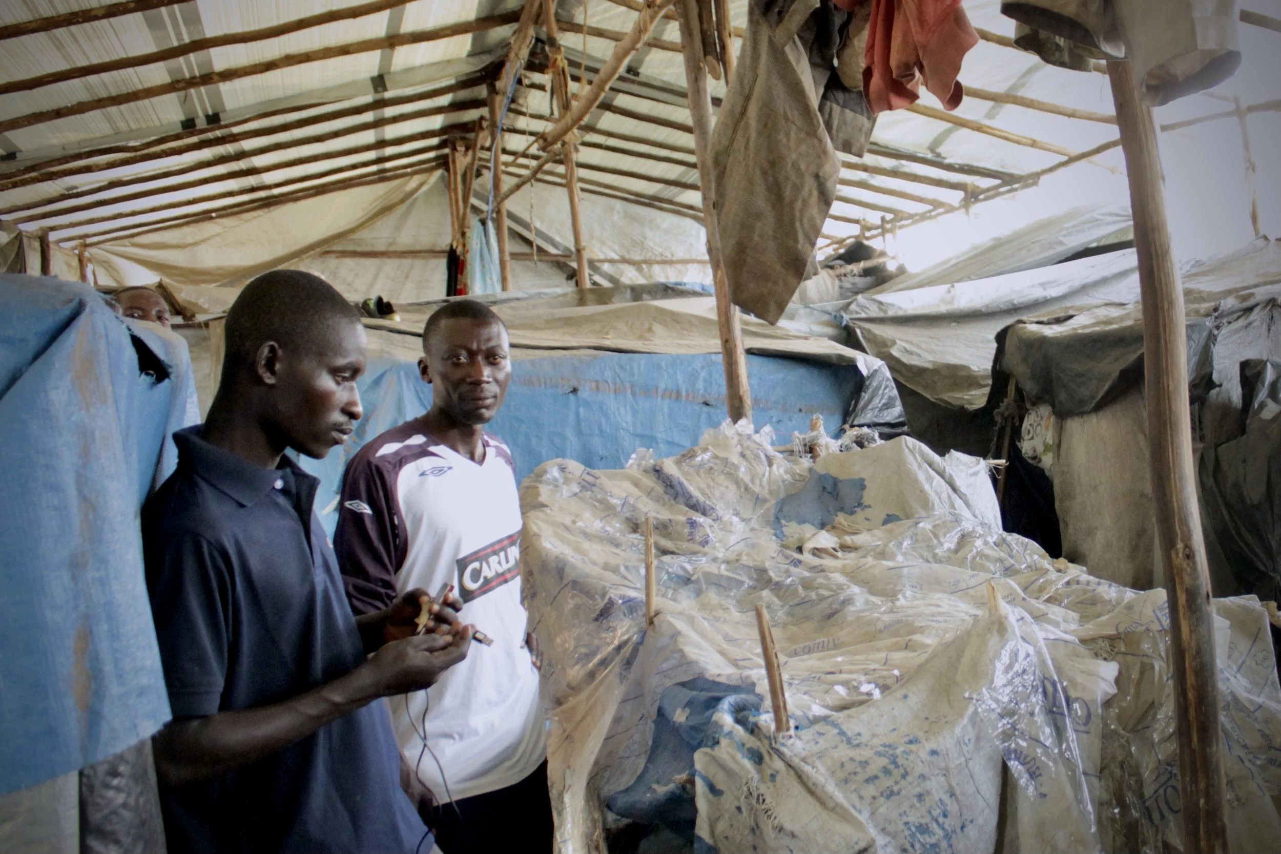 Two men, fathers, stand inside a temporary shelter, where plastic sheeting, wooden poles and hanging clothes formed cramped homes for displaced families.