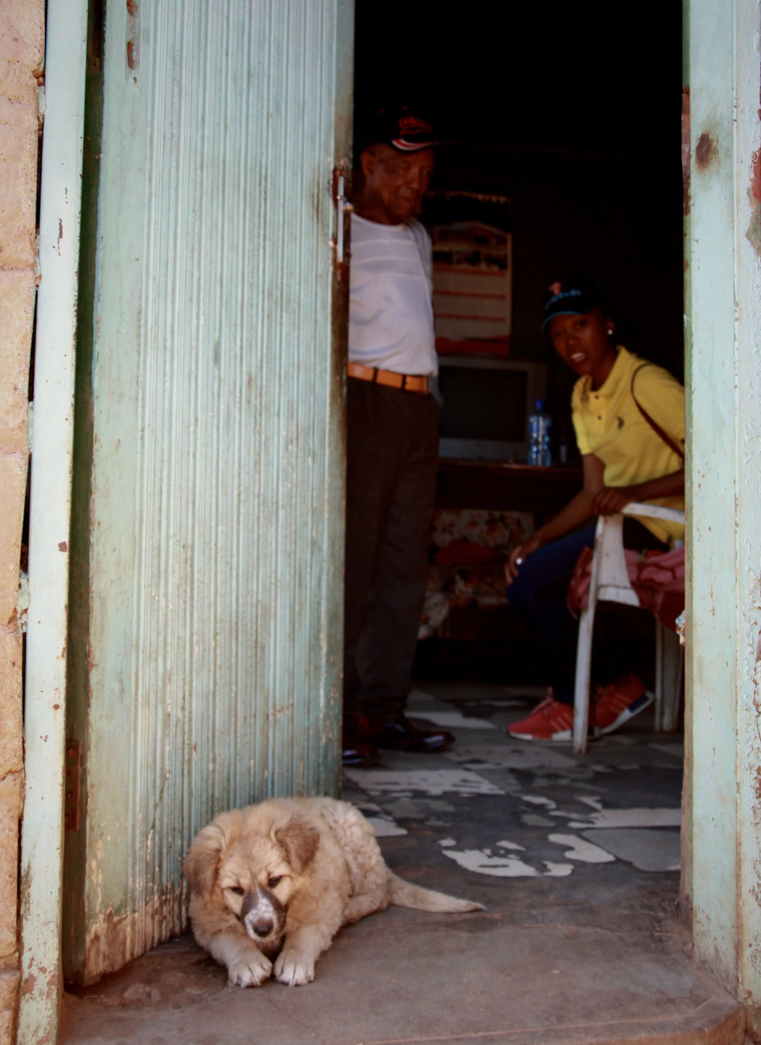 Viewed through the doorway of the family home, a dog rests on the floor while Michael and a visitor speak inside. The household was referred to Sentebale’s OVC programme through local community structures after village leaders identified the children