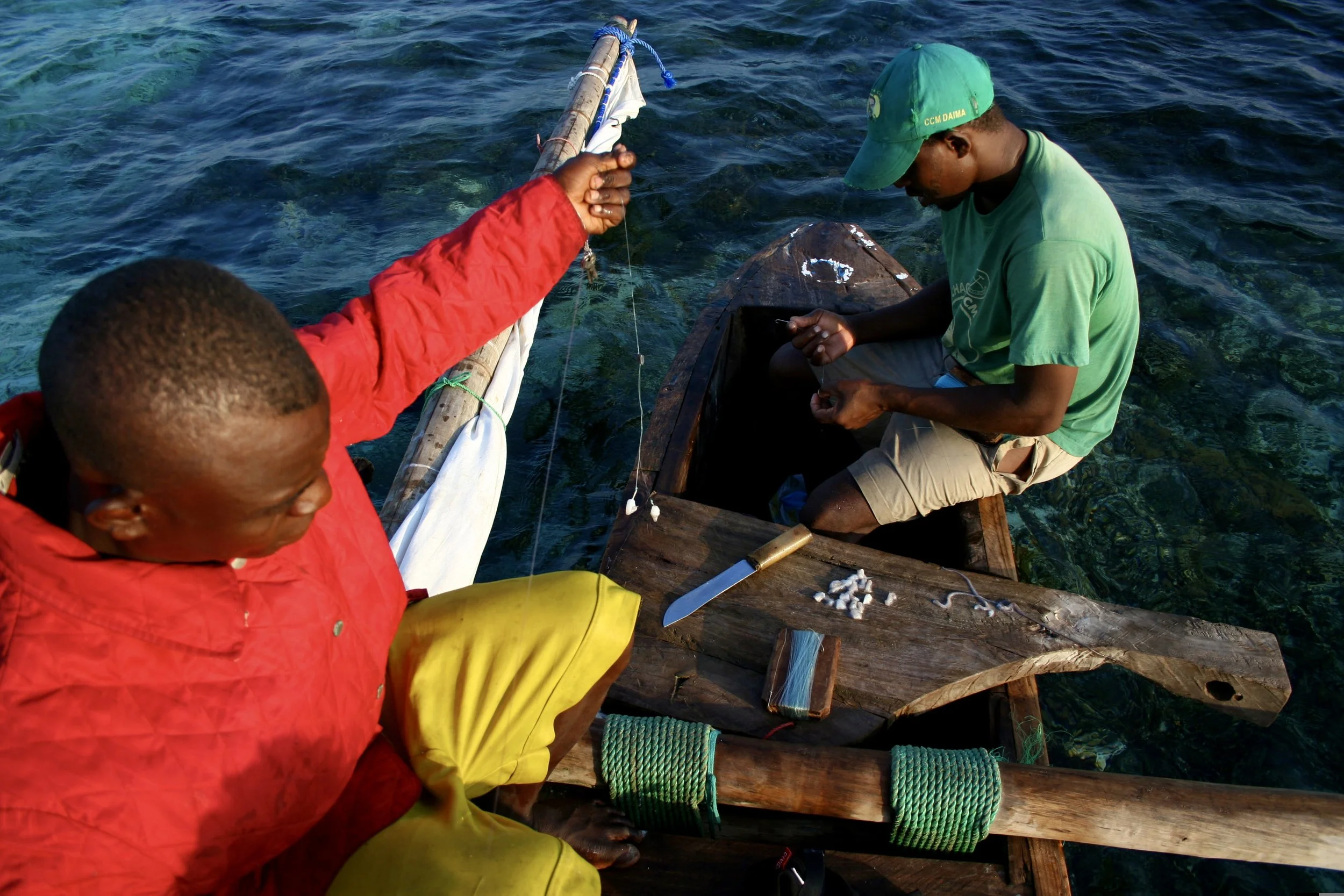 Crew members prepare hand lines and hooks aboard the dhow, knife and bait laid out on the worn timber gunwale.