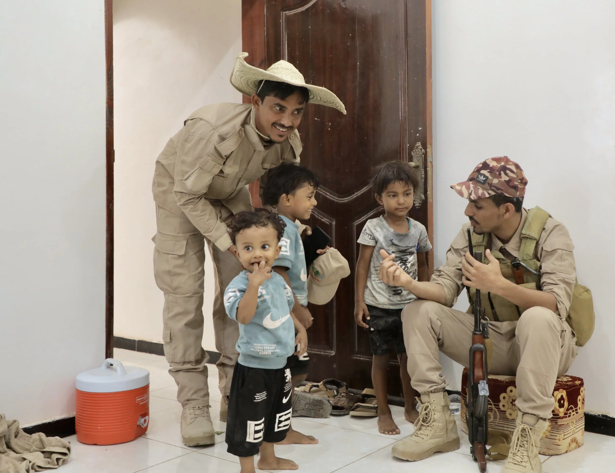 Sami returns home wearing a traditional handwoven straw hat after a day working in mine clearance. Armed guards assigned to the team relax alongside children inside the house.