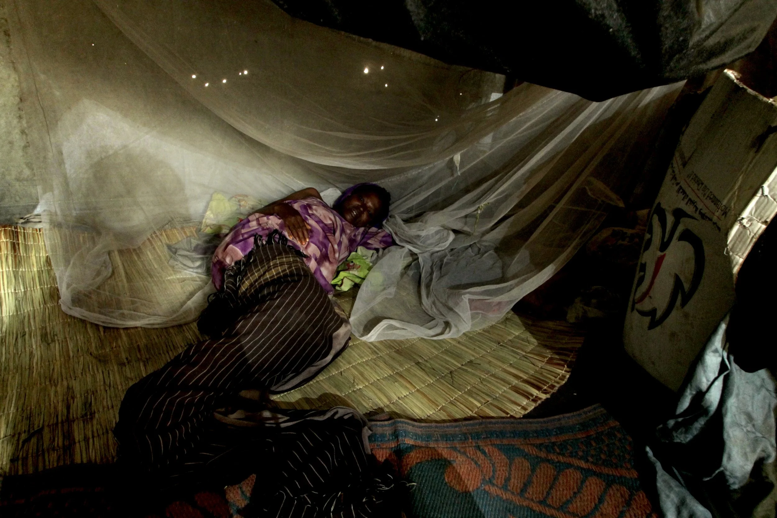 An ill woman lies beneath a mosquito net inside her shelter, a common protection against malaria in the crowded camp.