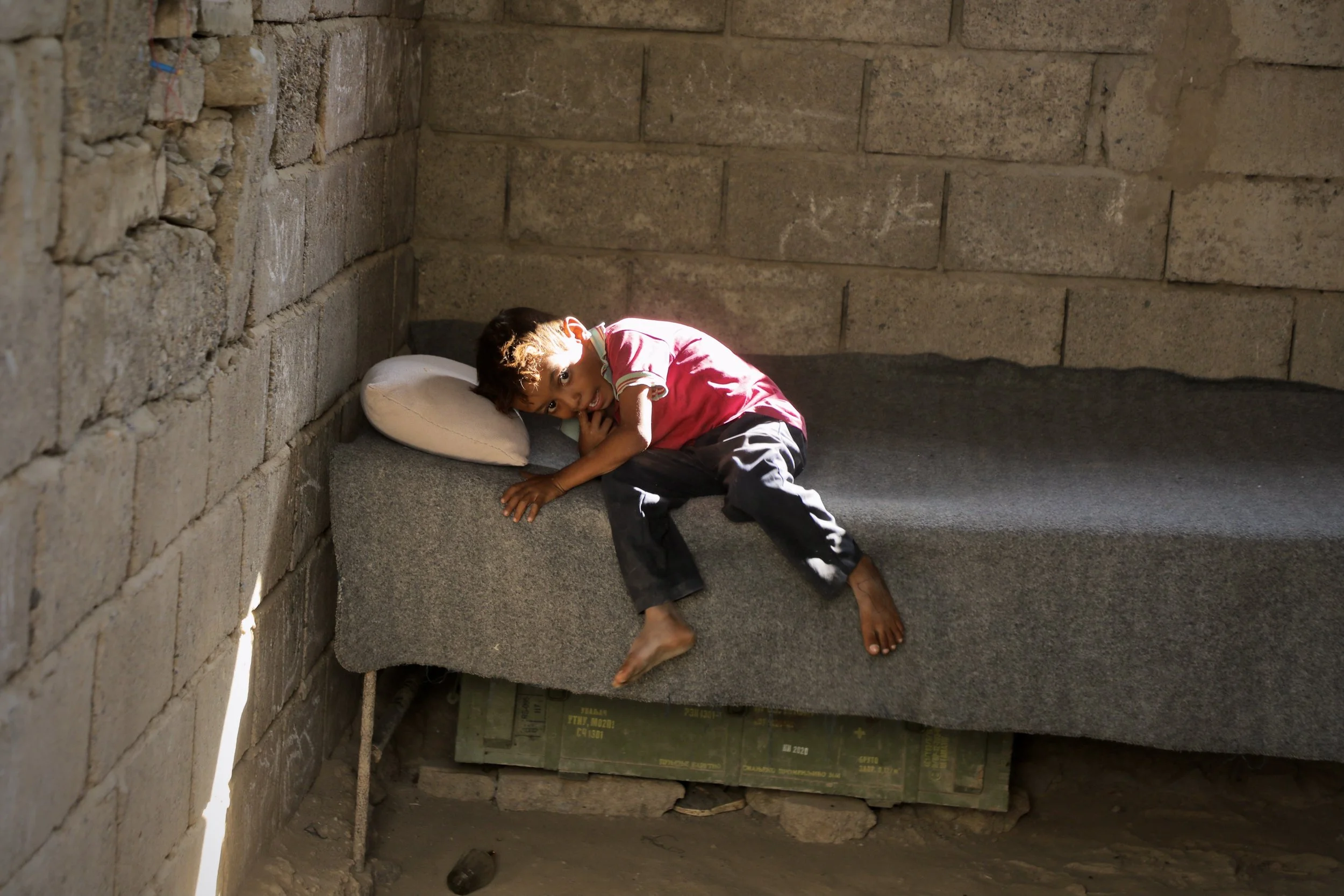 A child rests inside the family home. Ammunition crates repurposed beneath the bed remain a quiet trace of the conflict that once displaced the family.