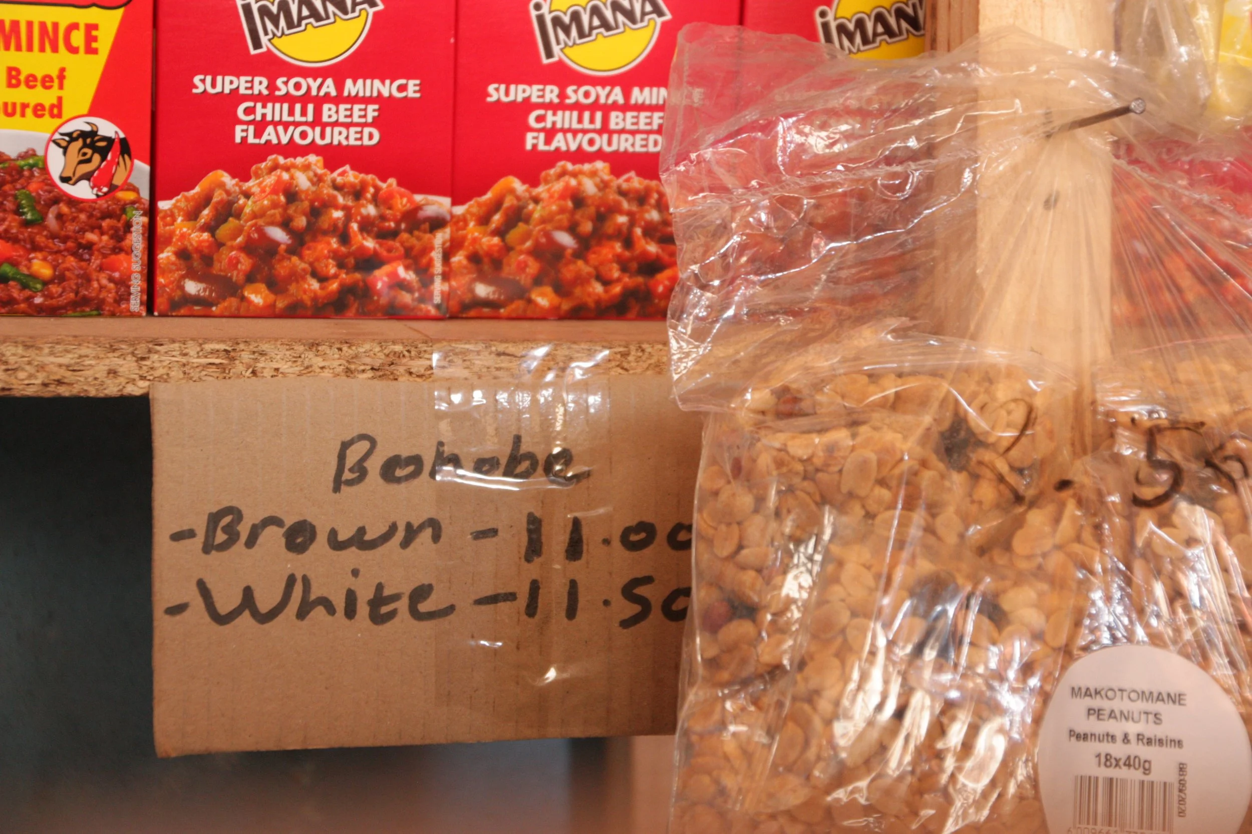 Handwritten prices for bread and packets of peanuts hang beside shelves stocked with inexpensive food items inside Paballo’s shop. Customers often buy goods in small amounts, reflecting the economic pressures many families face.