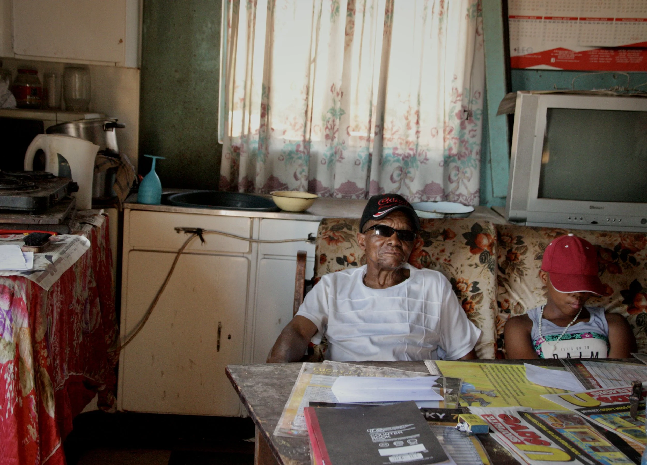 Michael Mosabala Leschibi, 78, sits beside his granddaughter Beauty inside the family’s modest home in Ha Chonapase, Leribe District, Lesotho. The family home reflects the financial pressures faced by many grandparents caring for vulnerable children 