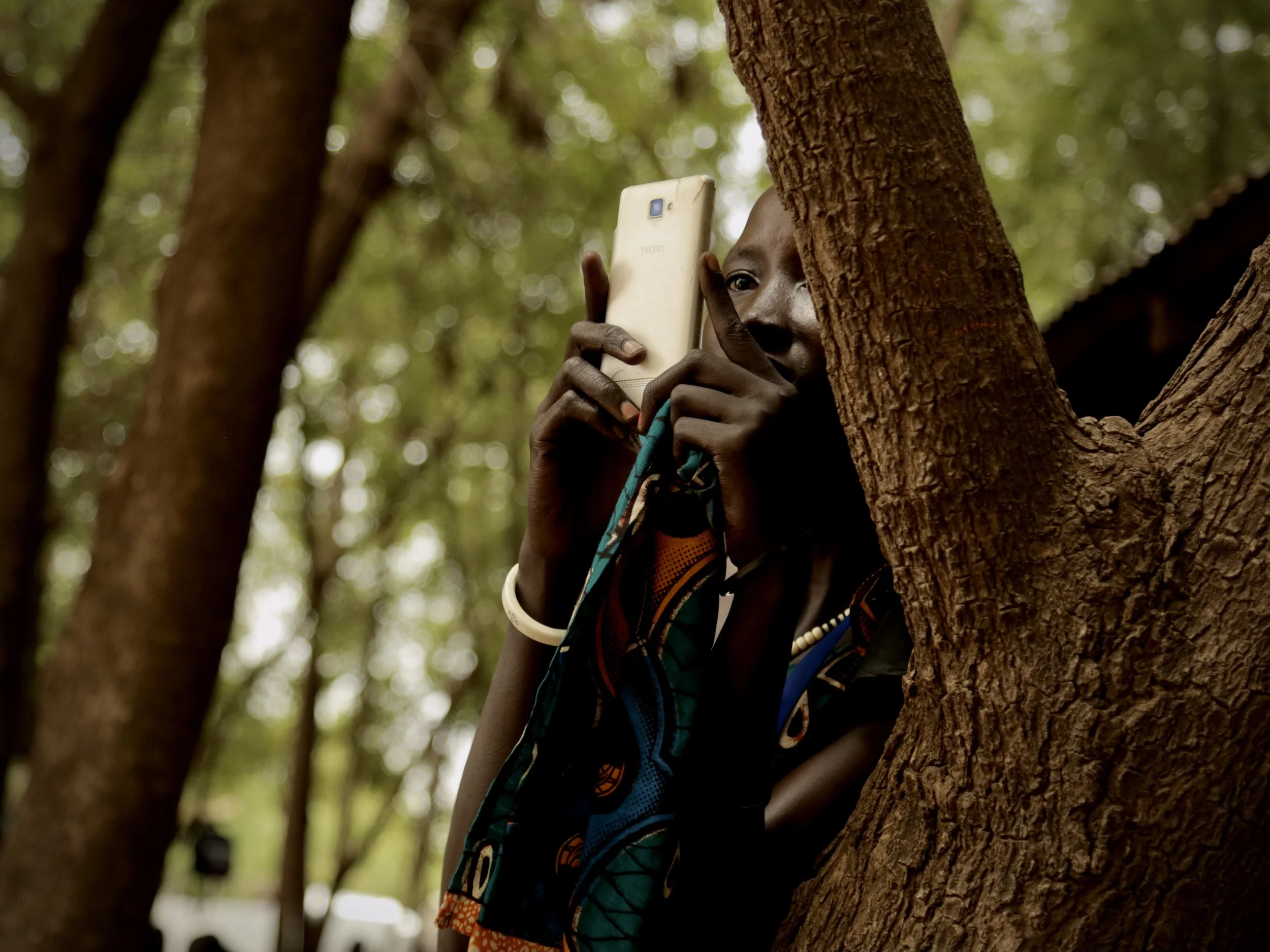 A girl photographs the scene while leaning through branches in Kakuma.