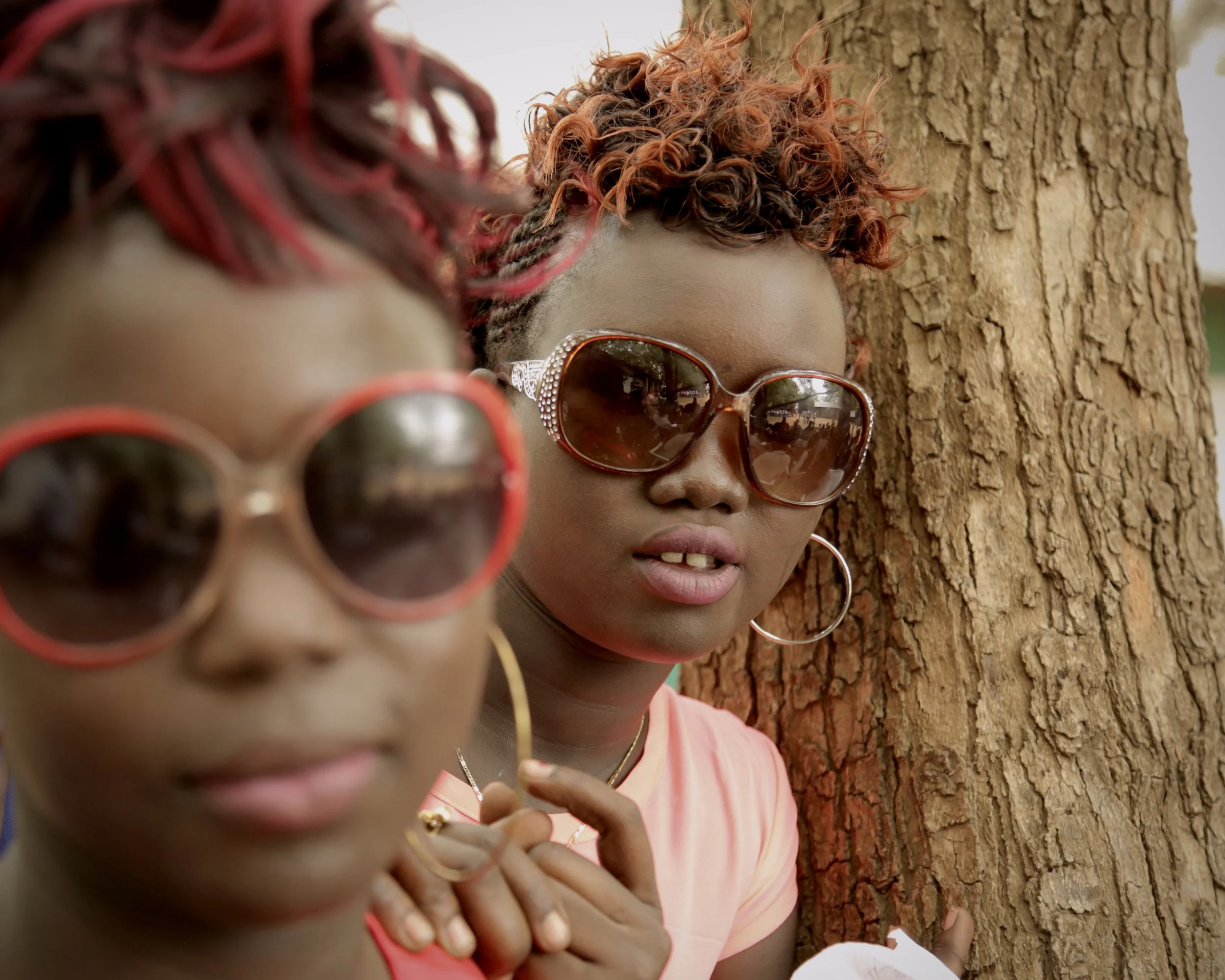Two young women pose beside a tree wearing sunglasses in Kakuma.