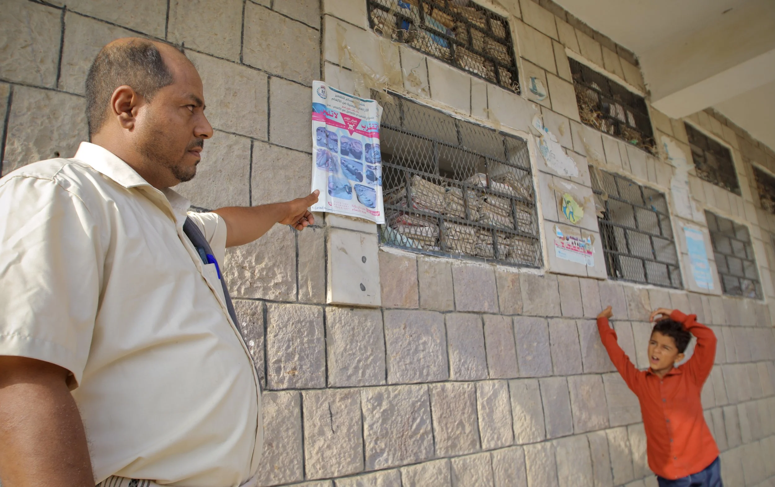 School Director  Ahmad Mohamad Al Barakani points to a mine-risk awareness poster displayed inside Al Shaab School while student Ahmed Mohammed stands nearby. Residents say children continue studying in partially damaged sections of the building.