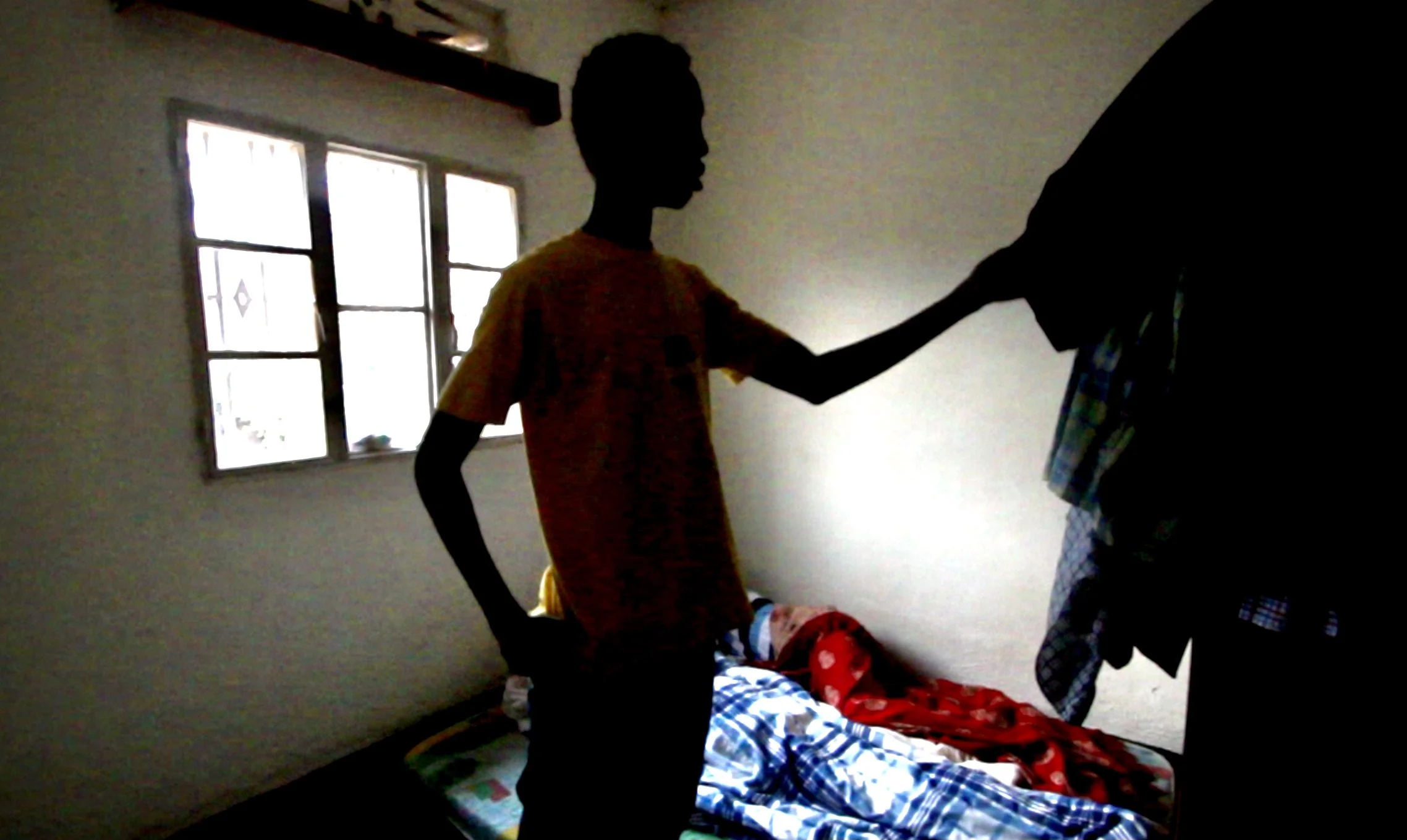 Justin Girukwigomba chooses clothes at daybreak inside the room he shared with other Burundian refugees in Kigali, Rwanda. Silhouettes of other residents remain asleep beneath blankets in the crowded room. Living with only a few personal possessions,