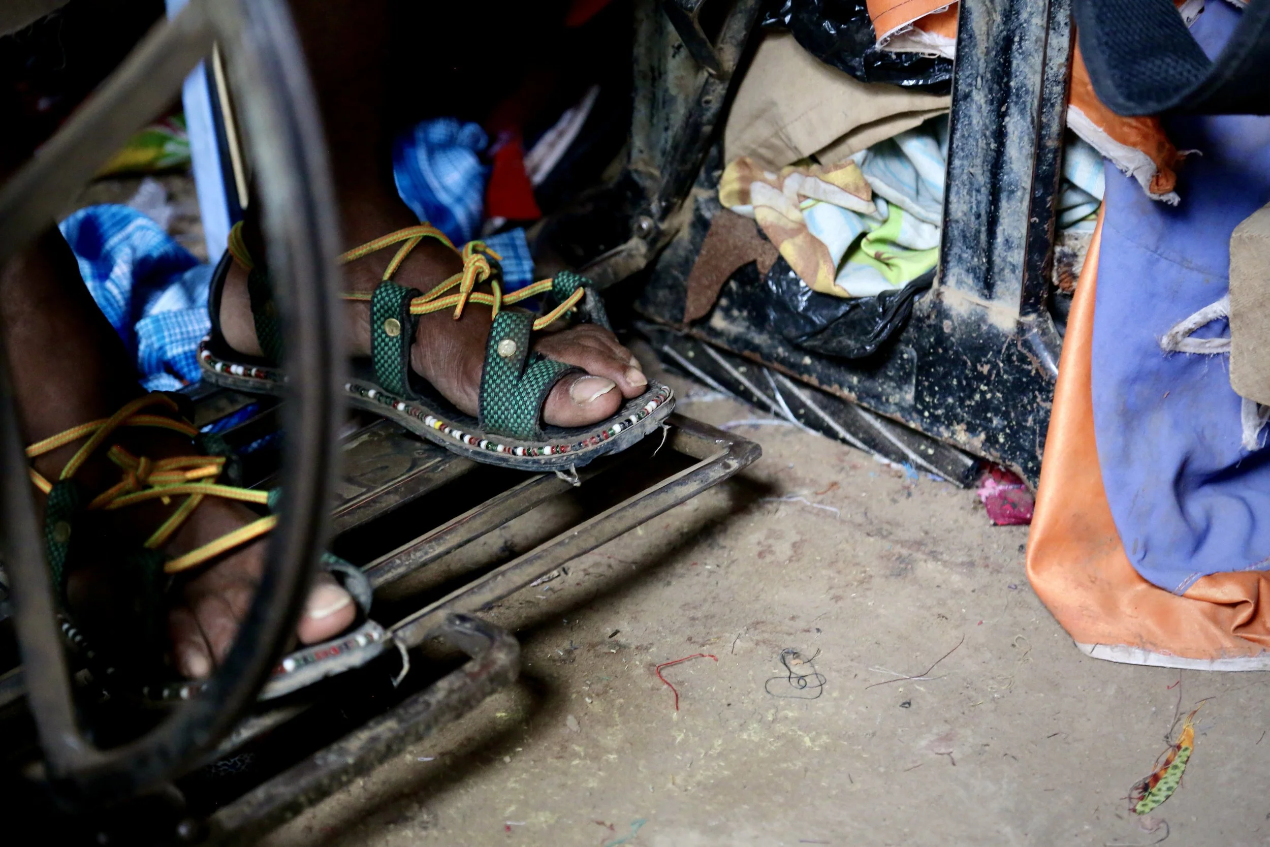 The tailor operates the foot pedal of his sewing machine. He wears locally made sandals, typical of the camp, crafted from recycled materials and decorated with beads.