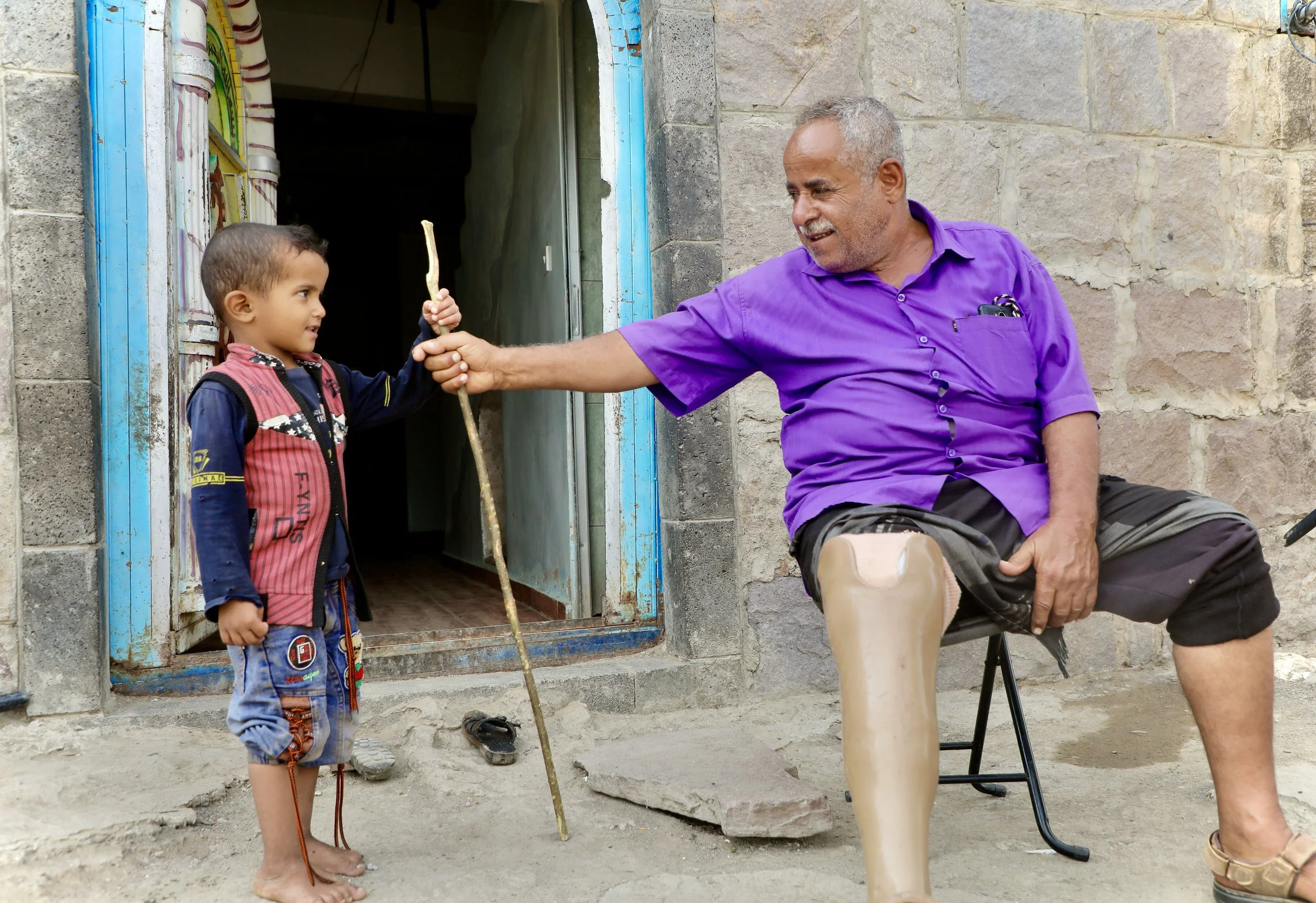 Mosaid Mohammed Ali Naji with his grandson outside their home in Shakhab, Al Dhale. His grandson holds out a stick - the gesture somewhere between an offering and a game. Mosaid's prosthetic leg is visible; he lost part of his leg when a landmine hid