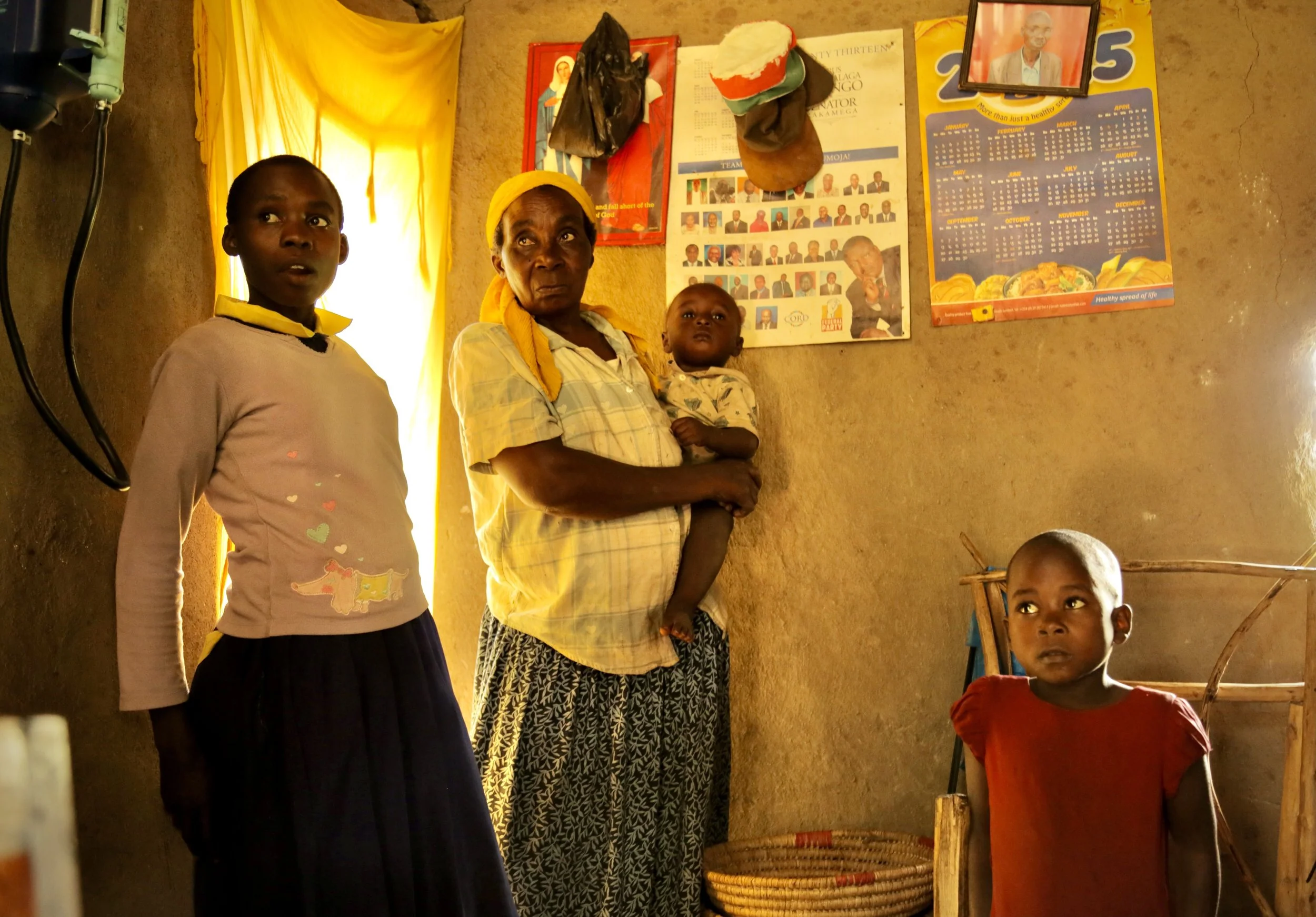 A grandmother stands inside her home with her grandchildren in Kakamega County, Kenya. Family members often play a central role in supporting children's education while managing household water needs.