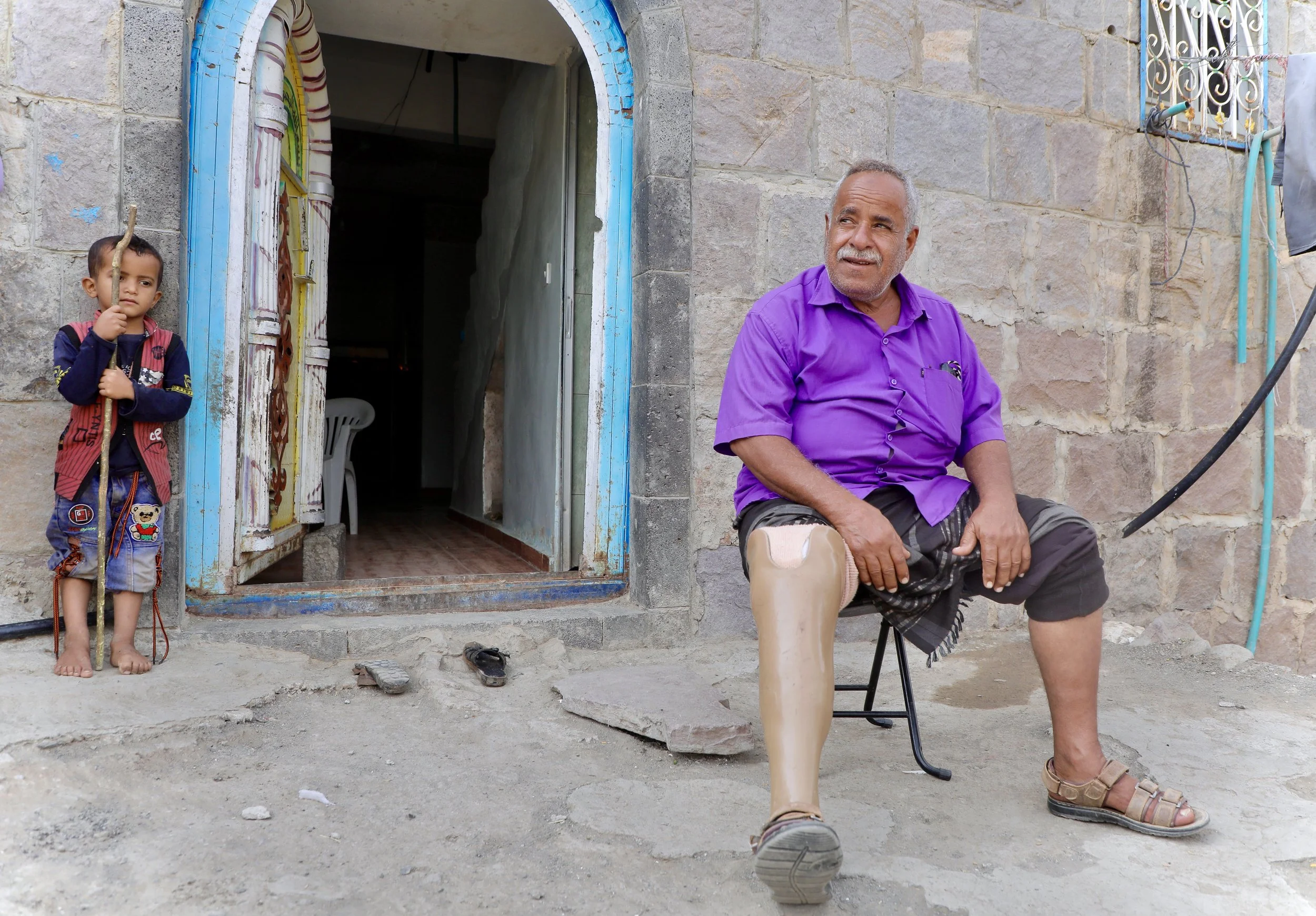 Mosaid Mohammed Ali Naji, Shakhab, Al Dhale. His grandson watches from the doorway behind him. When he returned home, he tragically discovered landmine and IEDs planted in this courtyard.