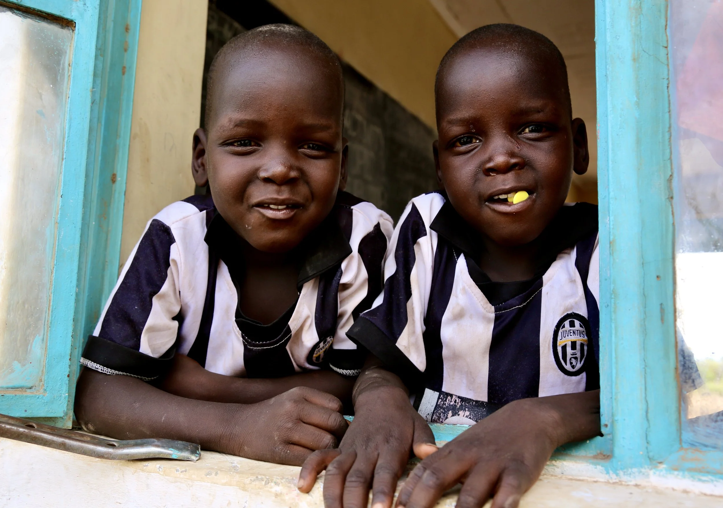 Two boys lean from a classroom window in matching Juventus shirts. Football kits travel farther than borders; in Kakuma, global teams become local uniforms of belonging, style and imagination.