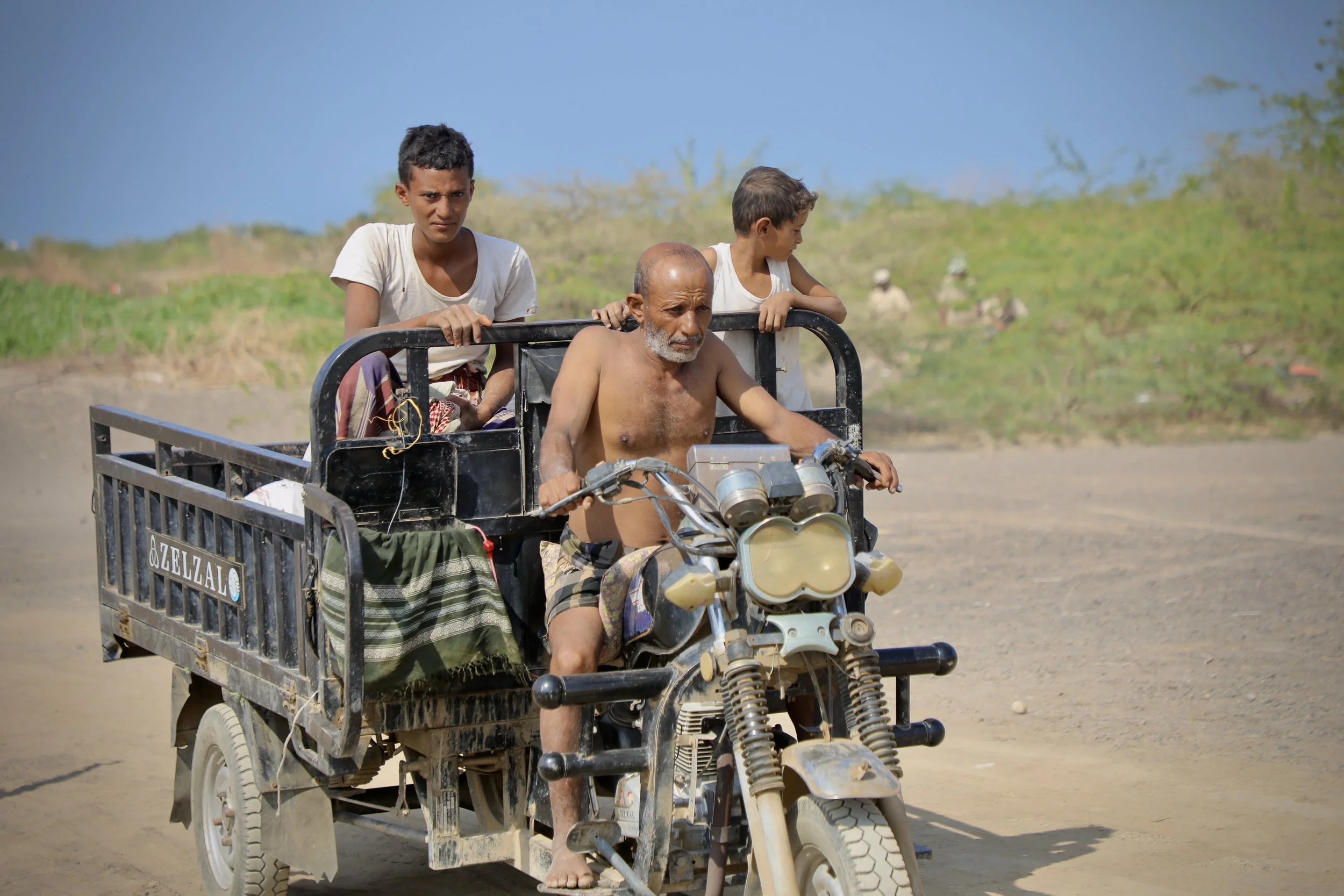 Men travel along a track while deminers work in the background in Al Remah, near Mocha, Taiz governorate, Yemen, where civilian movement and clearance activity often overlap.