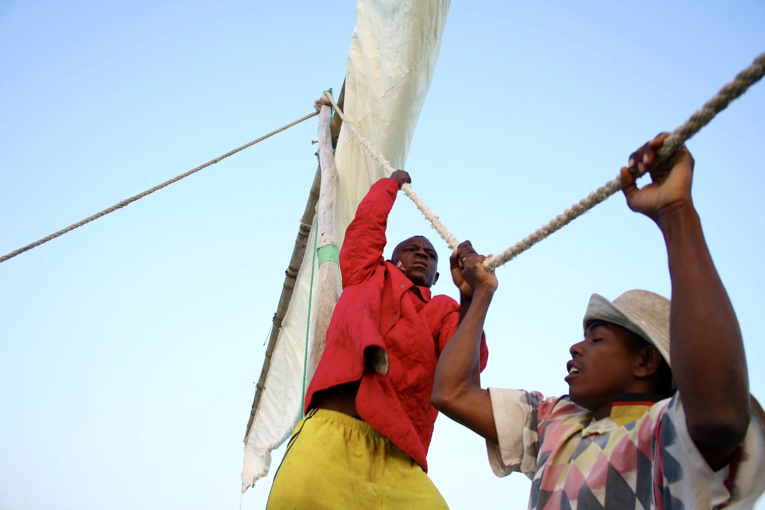 Two crew members pull together on the rigging — the physical choreography of raising a sail with rope and muscle rather than any mechanism.