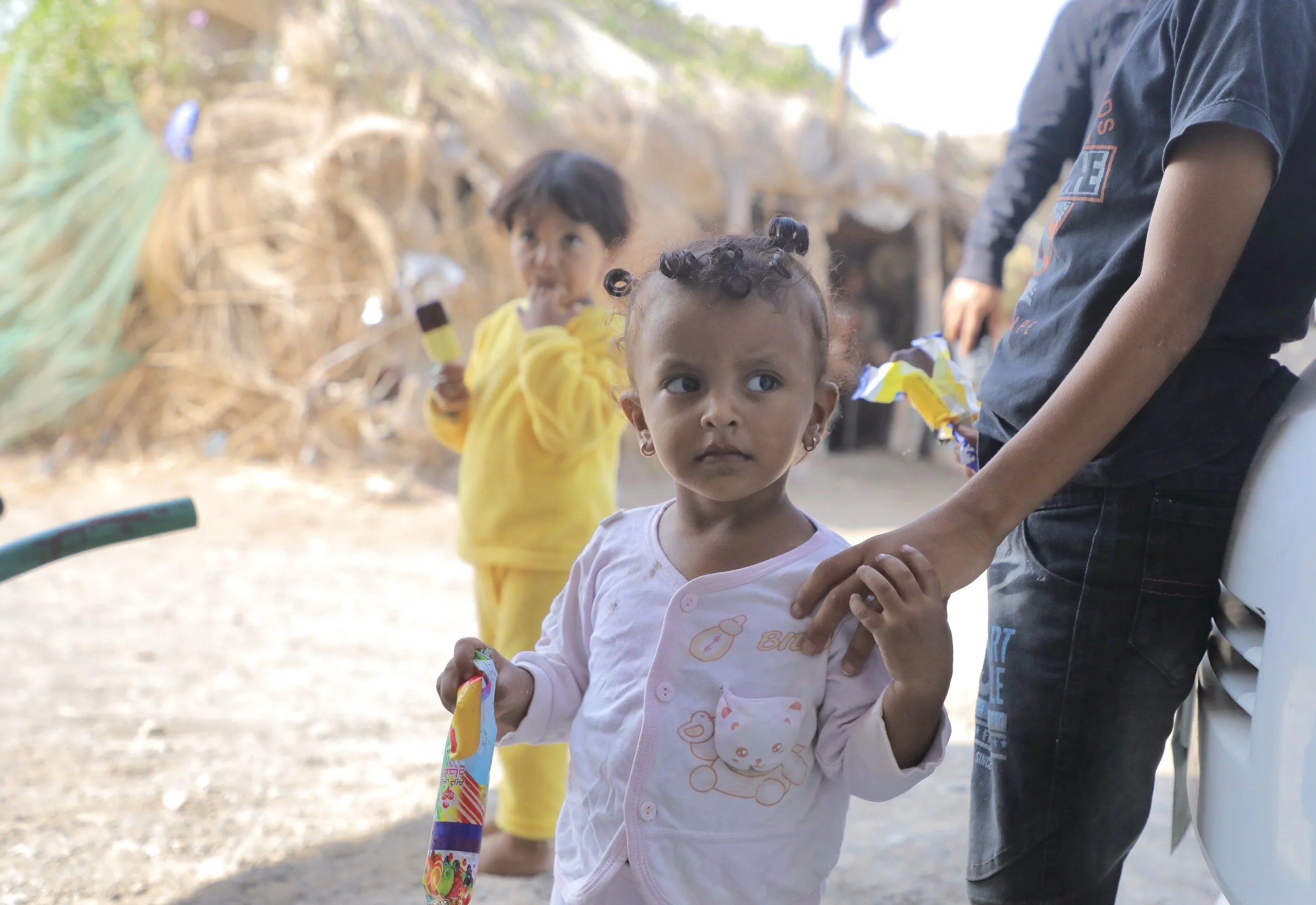 One of Sami’s young relatives, a niece, stands outside the family home holding an ice lolly as children move between neighbouring houses and the street.