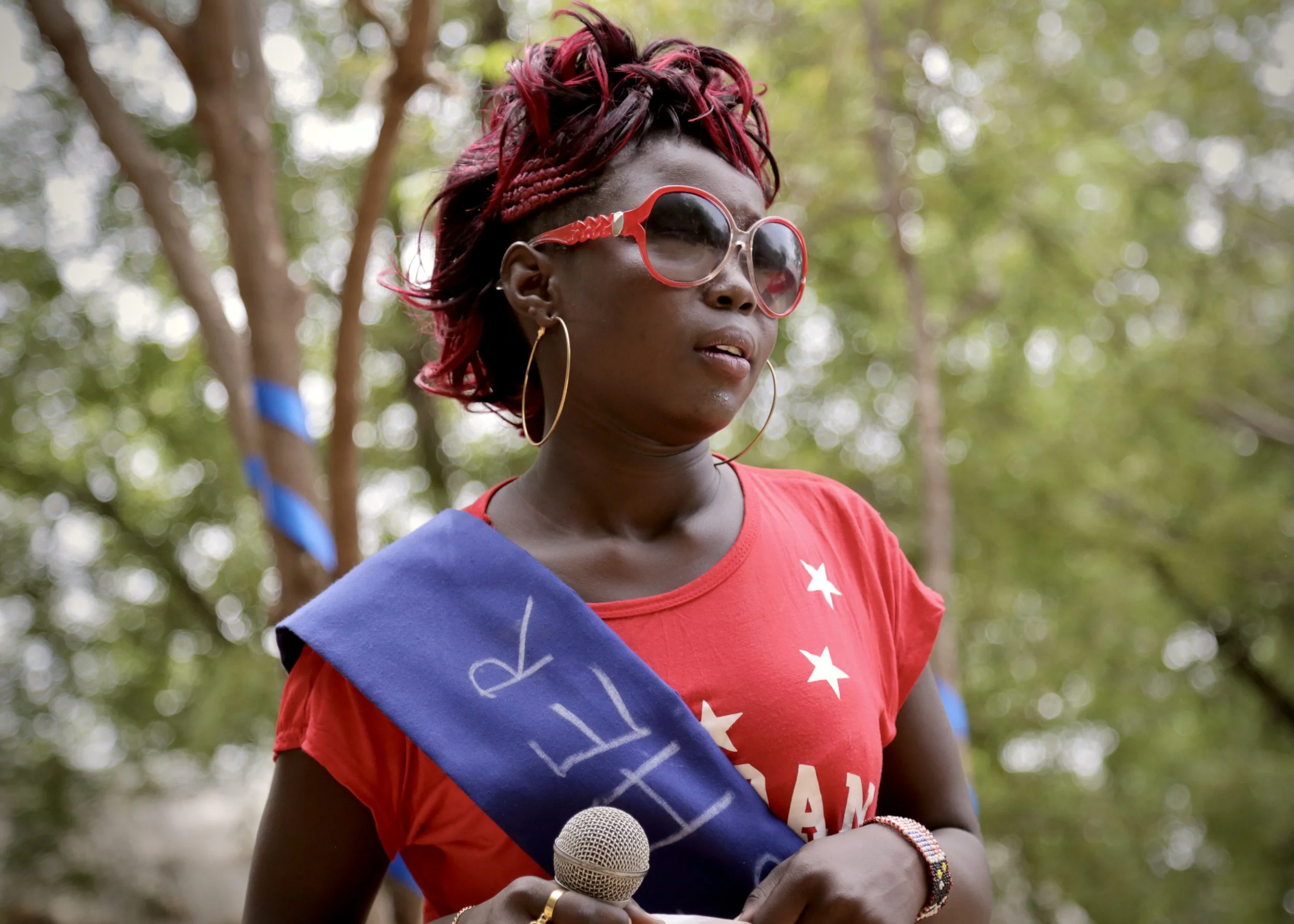 A young woman in a Miss sash and holding a microphone prepares to speak or perform in Kakuma.