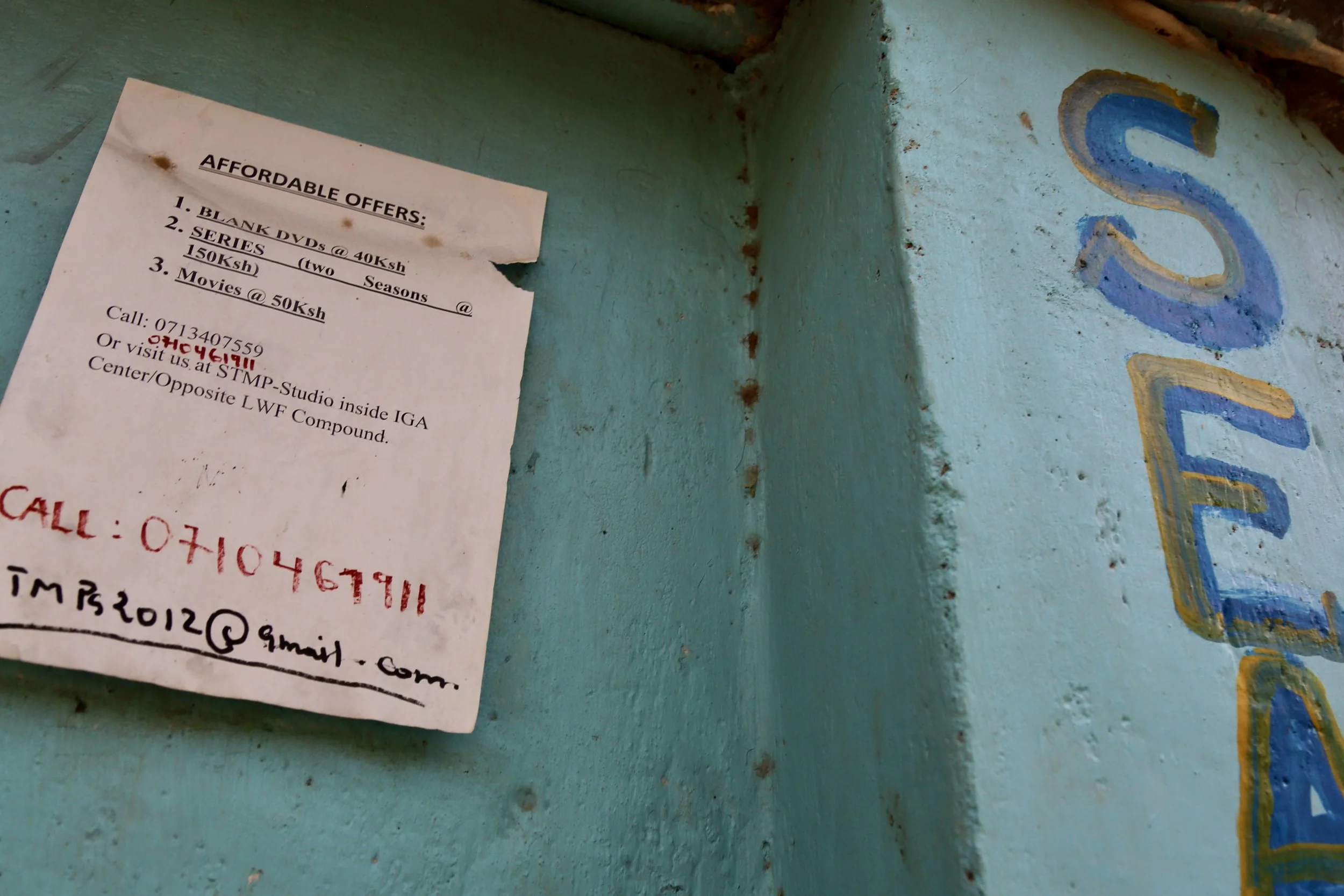 A price list and contact notice outside Fidele Wabenga’s studio in Kakuma Refugee Camp advertises DVDs, television series, and film services. The handwritten updates reflect the practical realities of running a small media business inside the camp.