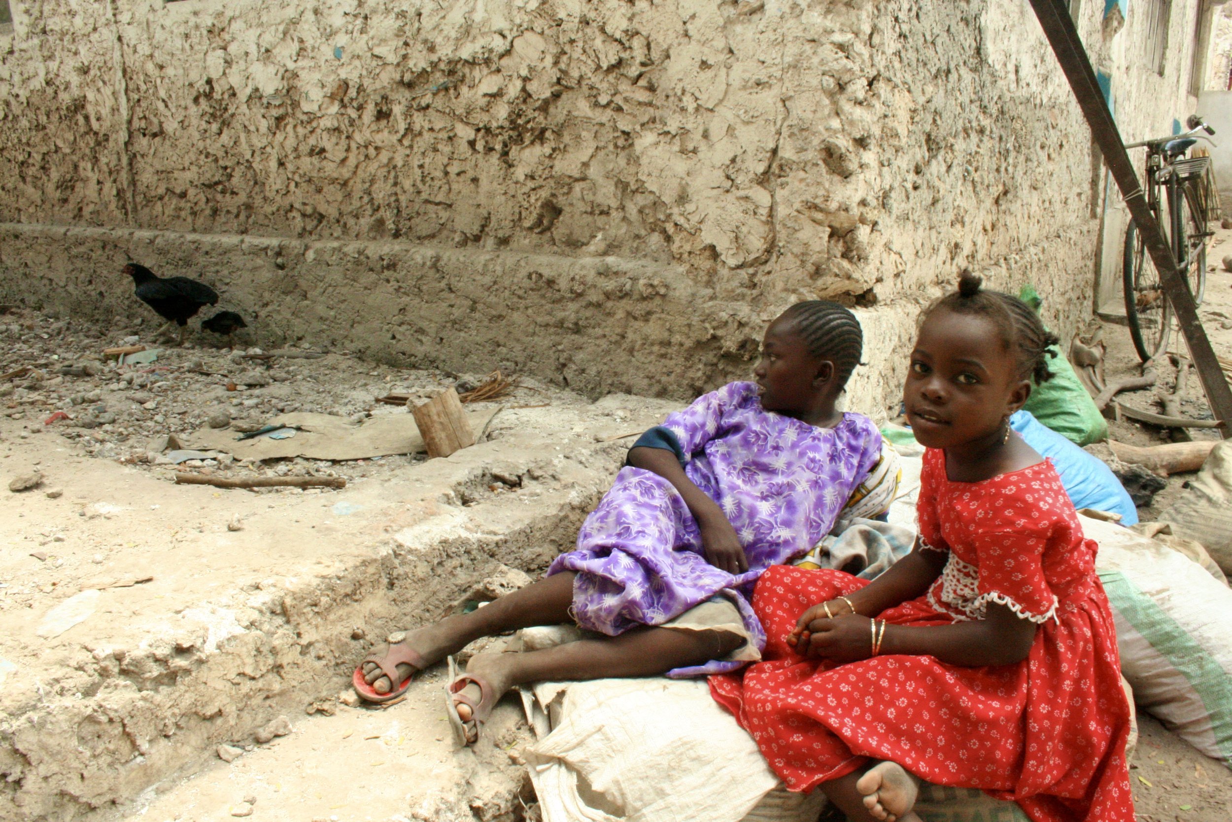 Two girls sit in a coral-stone courtyard elsewhere in the village. A closing note on the community this catch will feed, beyond the boat and beyond Captain Fruit himself.