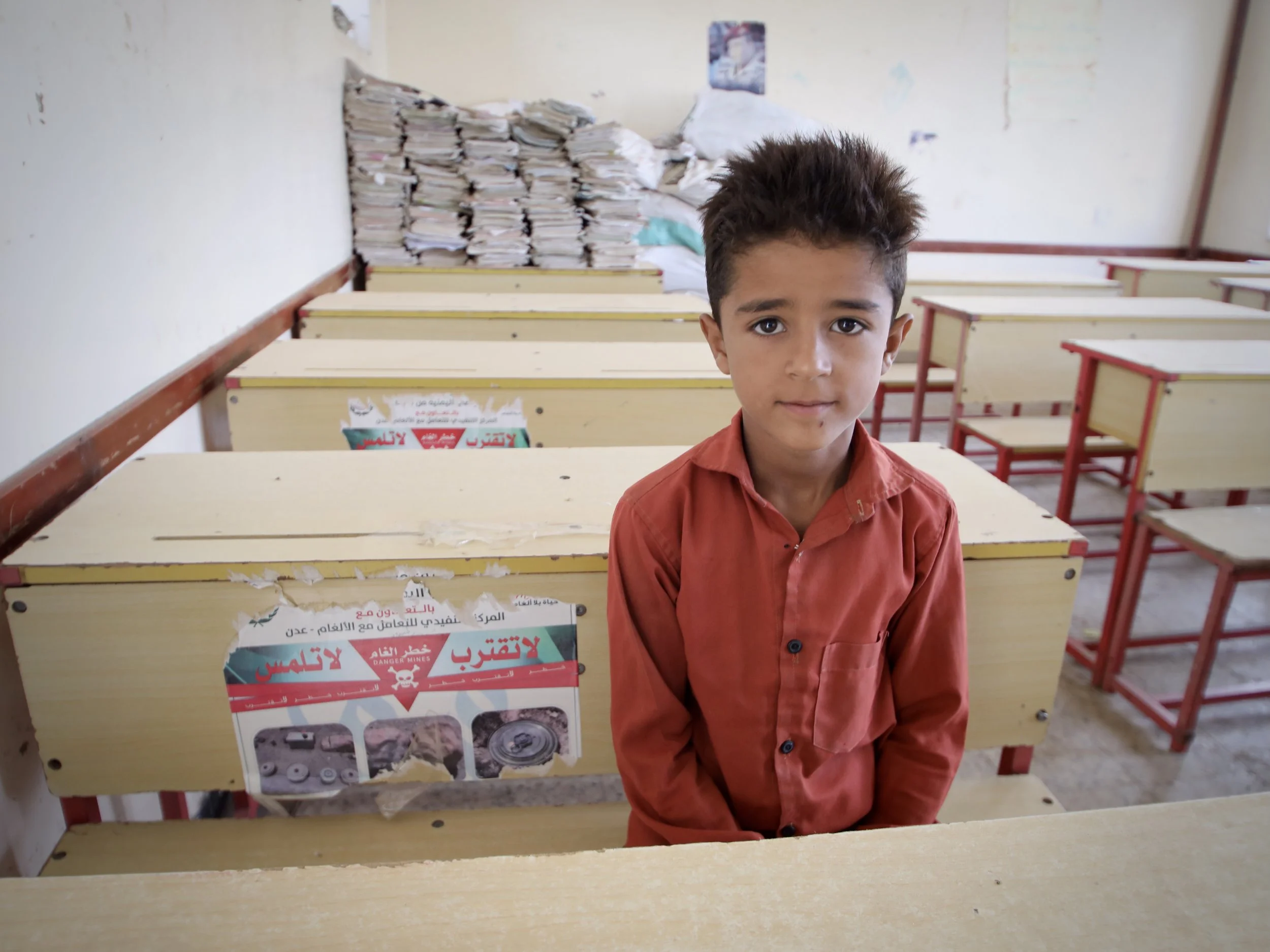 Marwan stands inside a classroom at Al Shaab School, where mine-risk awareness posters have been attached to school desks.
