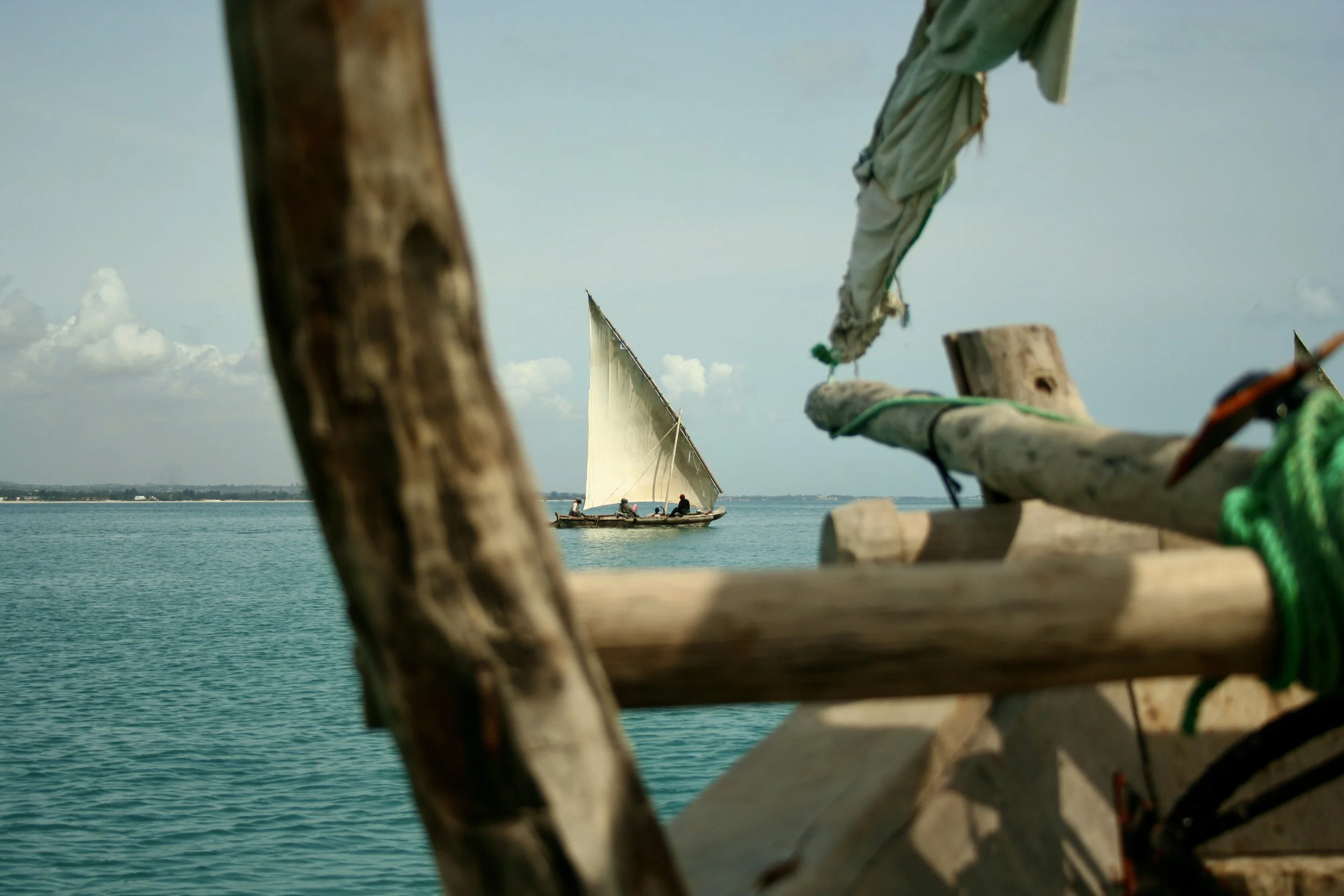 The dhow heads out from shore, seen through the rigging of a second boat; one working vessel framing another.