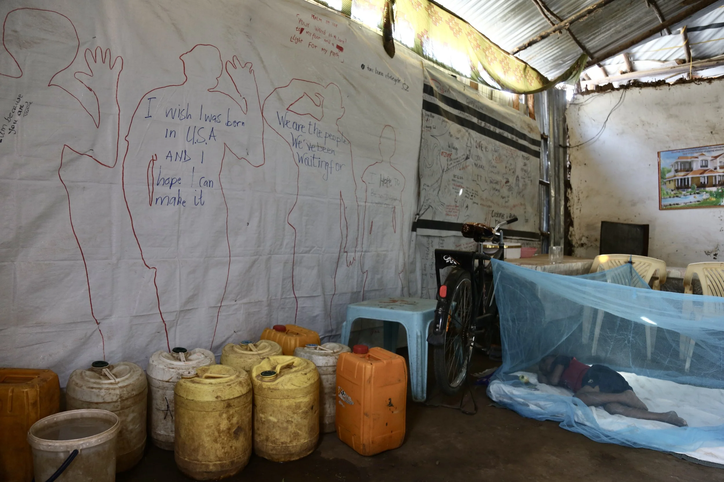 Inside the family shelter, handwritten messages cover the walls while a younger sibling sleeps beneath a mosquito net. The room holds both hardship and imagination.