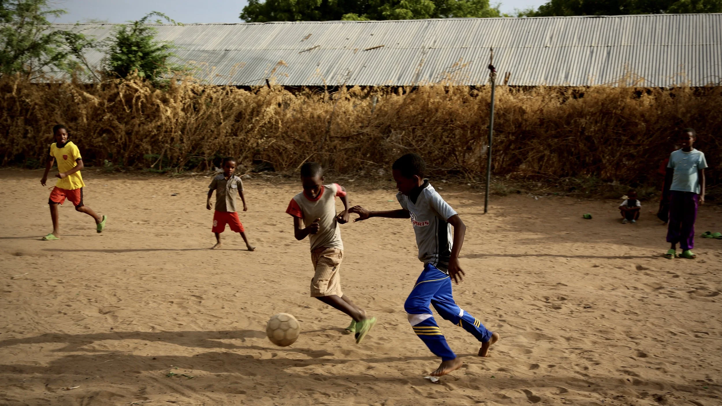 Children play football on a sandy pitch between shelters in Kakuma.