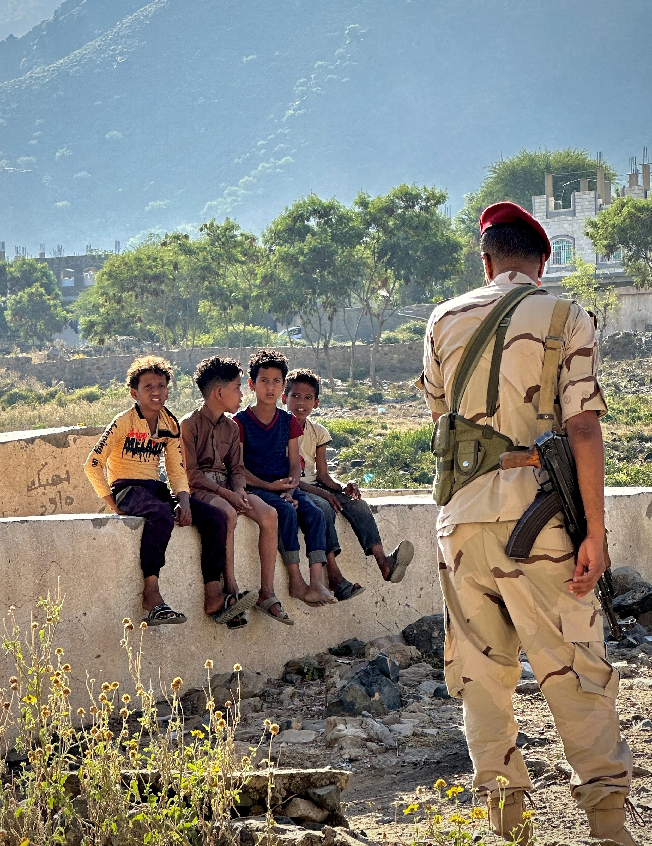 Children sit near an armed guard in the Ibrahim Aqeel School courtyard in Taiz. The school continues operating amid the long-term effects of conflict and landmine contamination in the area.