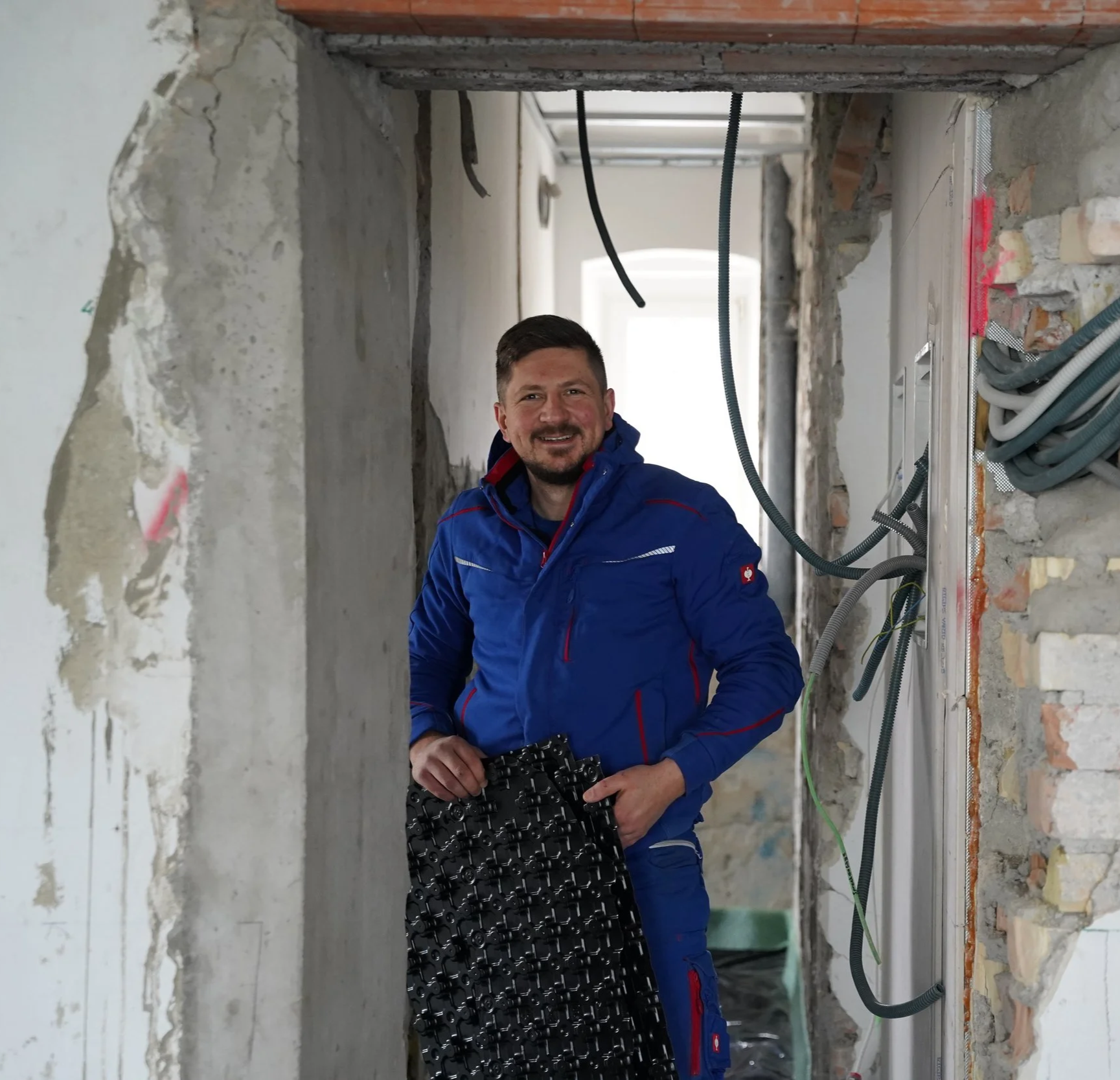 Ein Mann in blauer Jacke steht in einem Bauzimmer mit sichtbaren Rohren und Kabeln, hält einen schwarzen Bauwerkzeug und lächelt in die Kamera.
