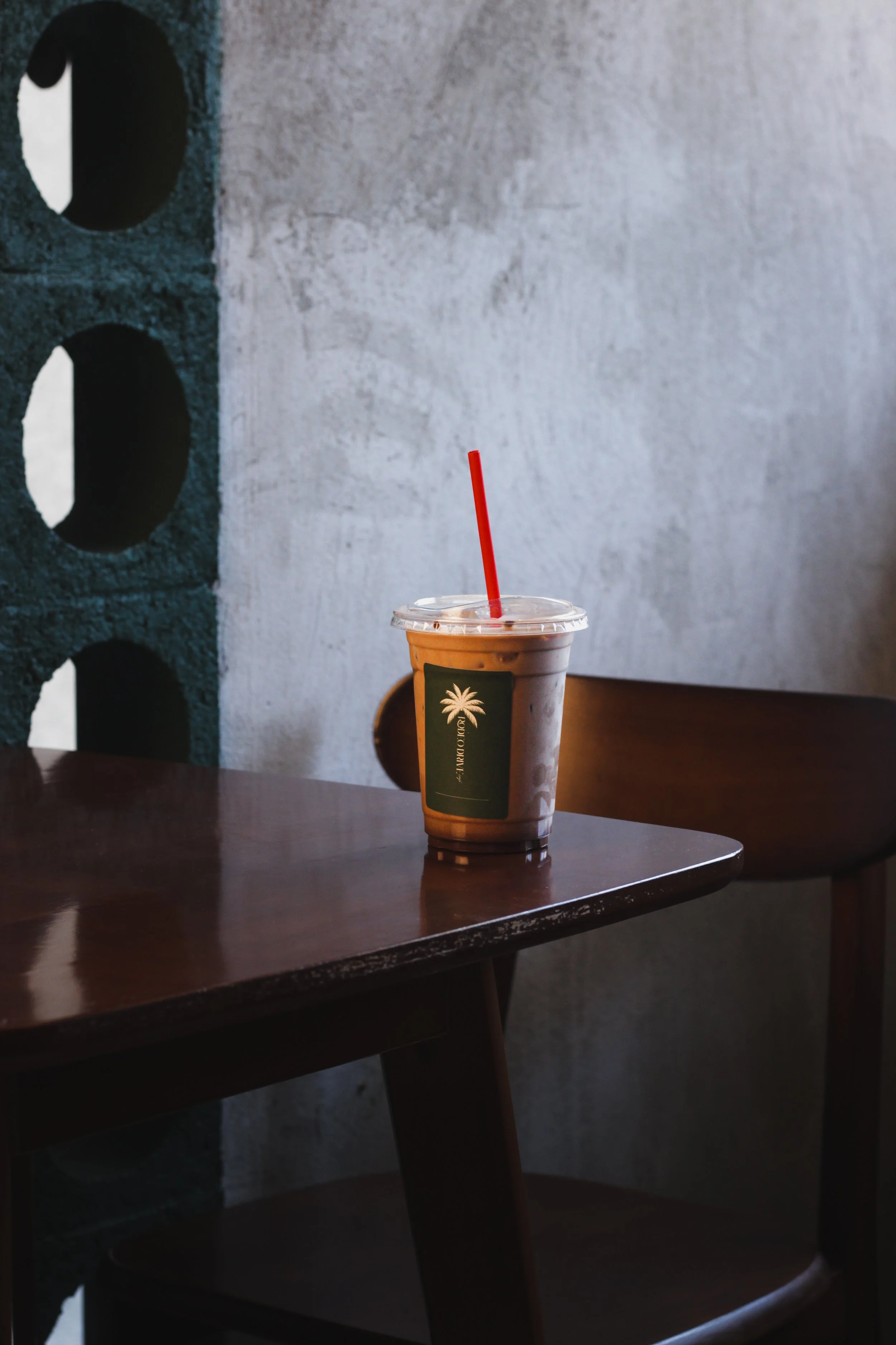 A plastic cup filled with a beverage, possibly iced coffee or tea, with a red straw and a lid, placed on a wooden table in a cafe or restaurant.