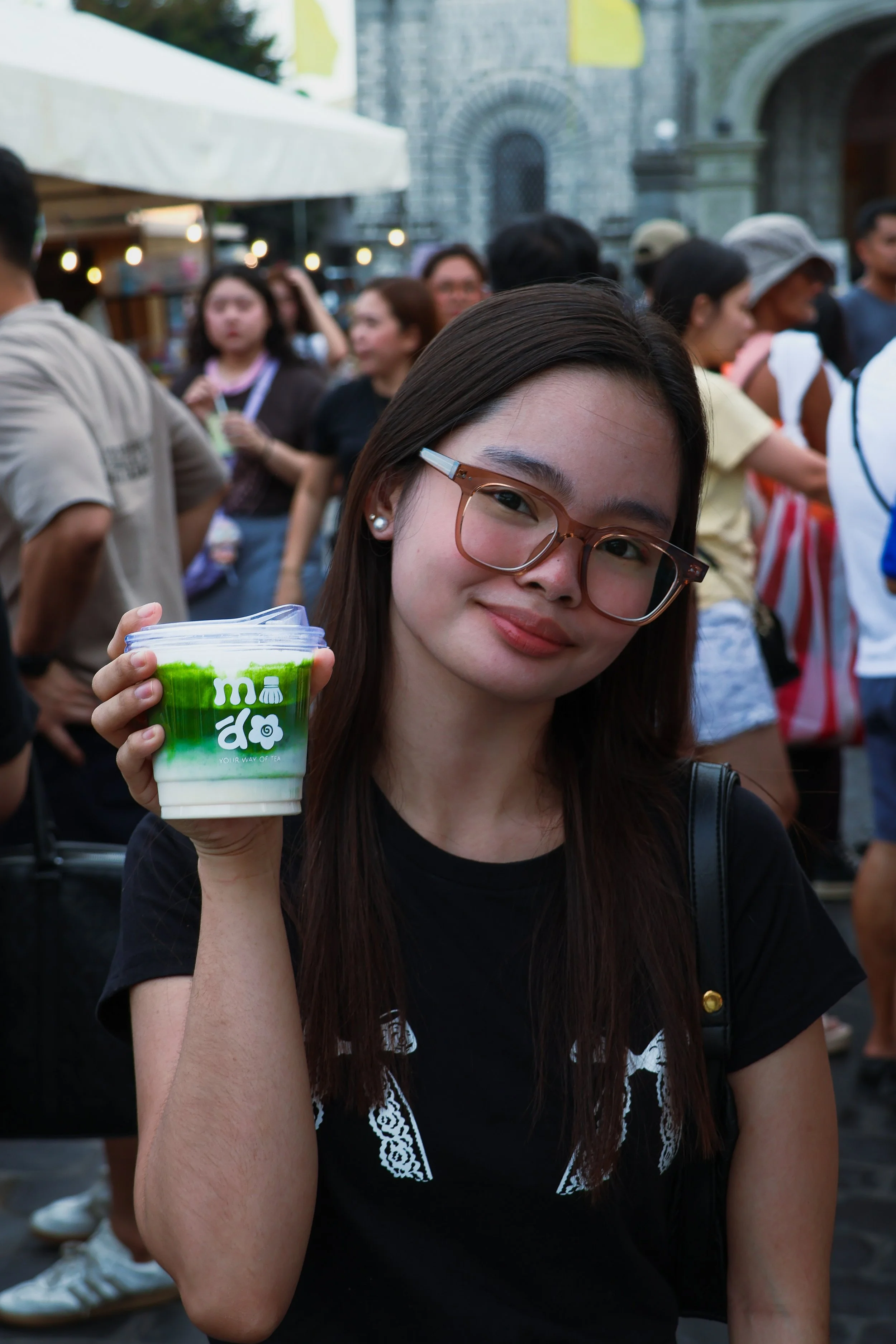 A young woman with glasses and long brown hair holding a cup of green and white drink, smiling outdoors at a crowded event with people in the background.