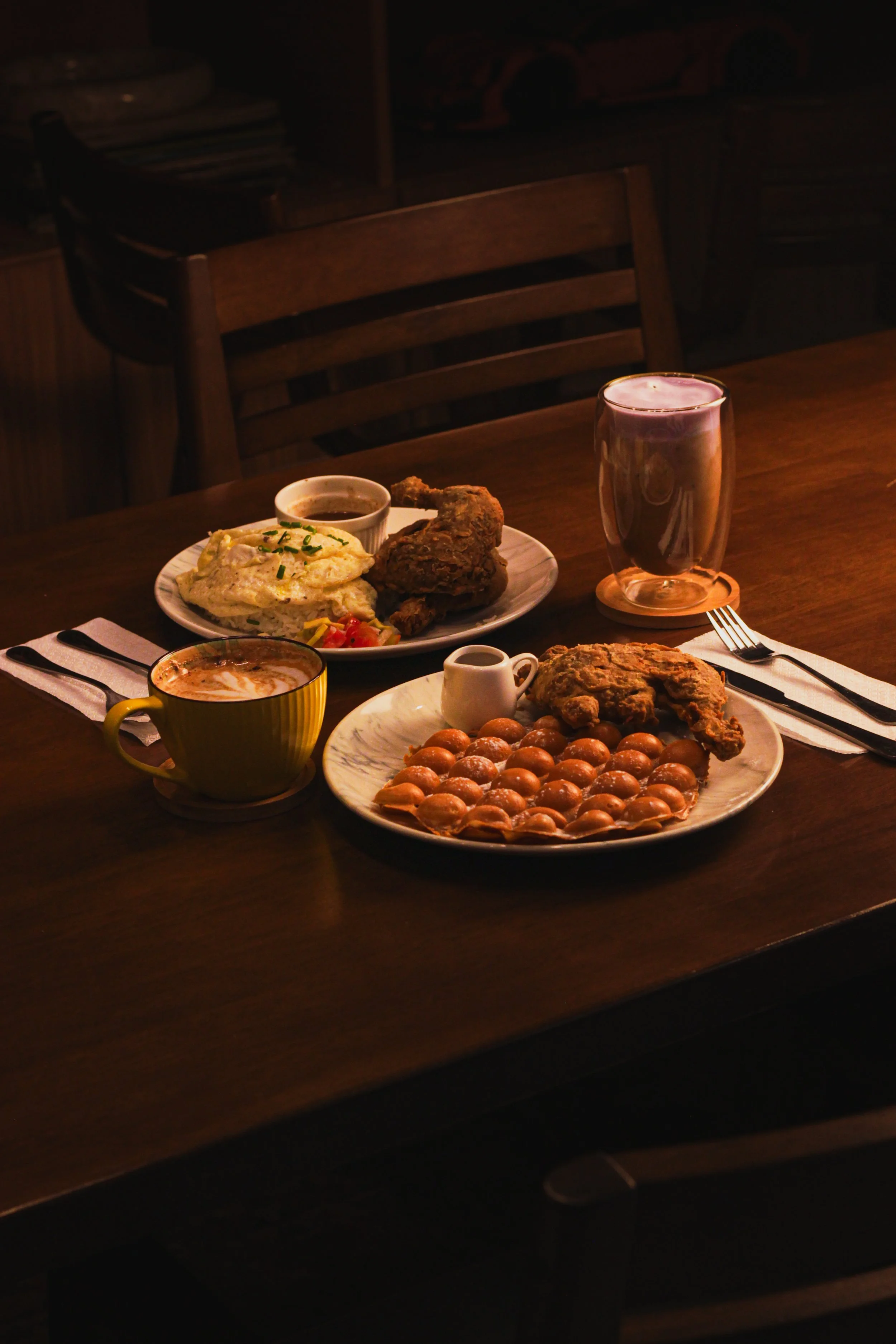 A wooden table with various breakfast foods and drinks, including fried chicken, scrambled eggs with chives, a small bowl of syrup, a glass of dark soda, a coffee mug, and a plate of round fried items.