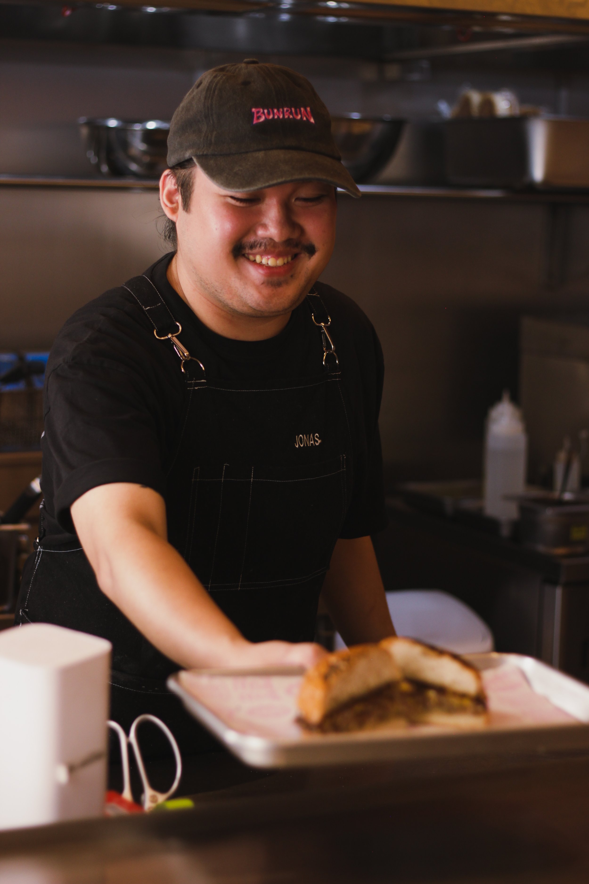 A smiling man wearing a black cap with pink text and a black apron working in a kitchen, with a plate of food in front of him.