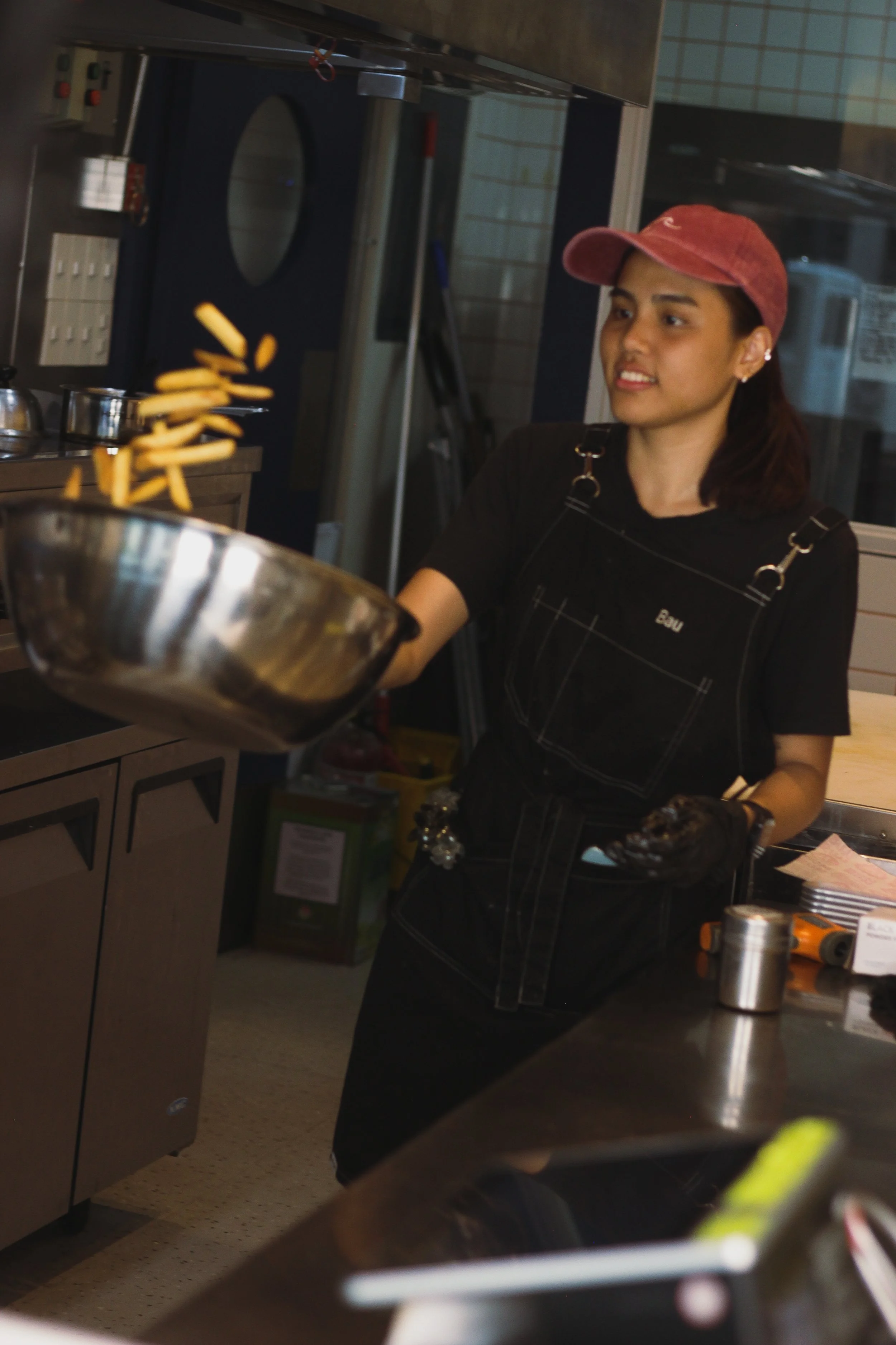 A woman wearing a red hat and black apron is cooking French fries in a commercial kitchen.