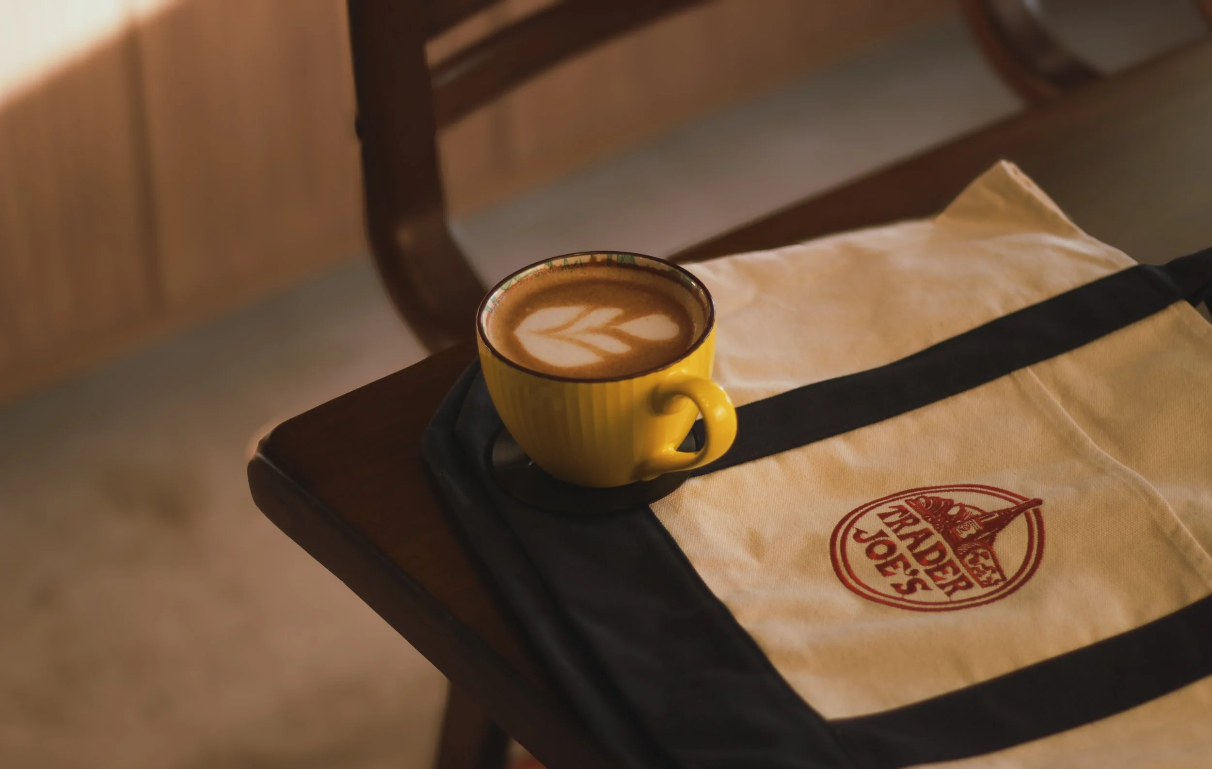 A yellow ceramic mug with a latte art leaf on top, placed on a black tray on a wooden table, next to a white paper napkin with red and black logo.