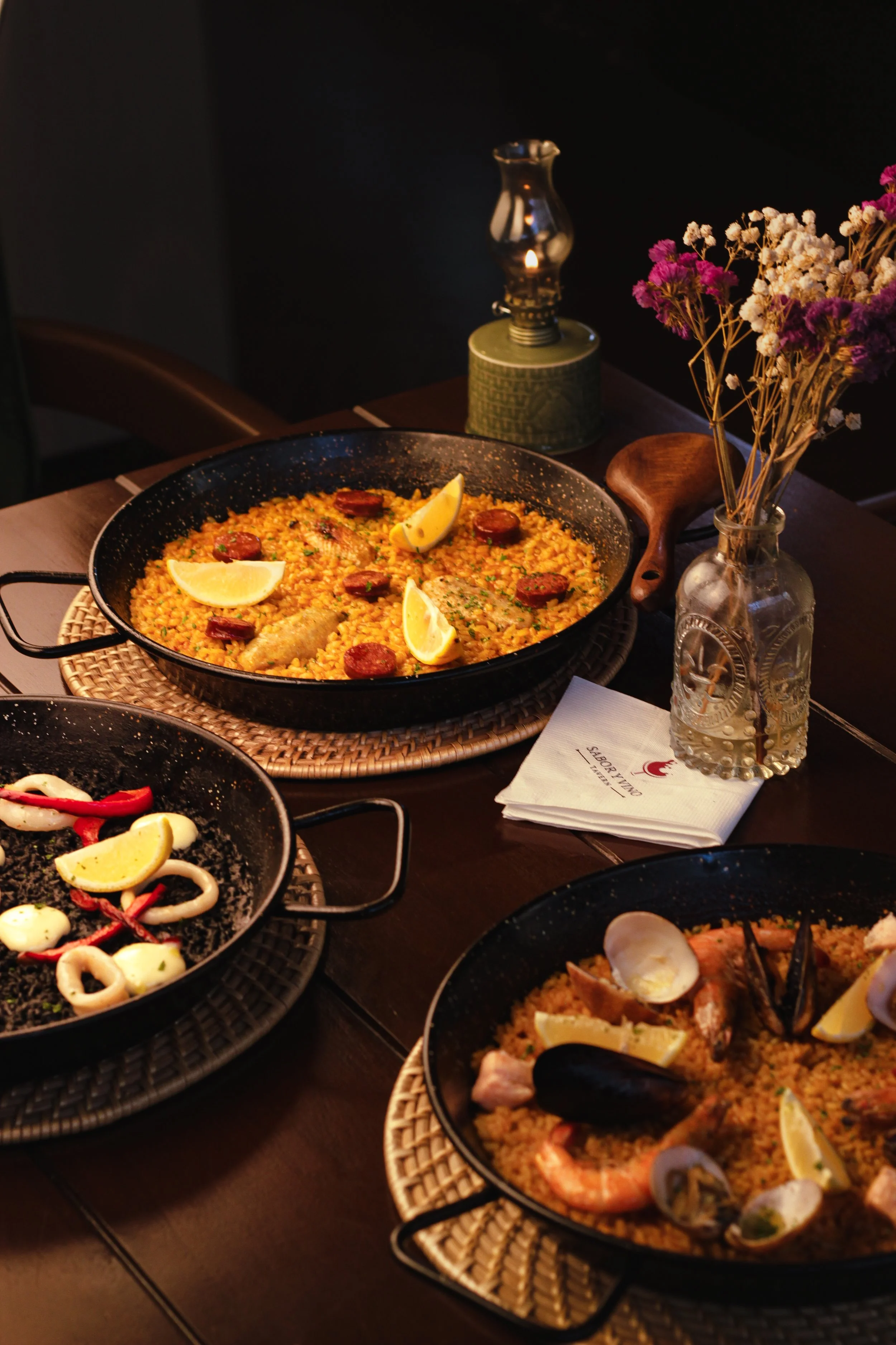The image shows a table with three black pans of Spanish paella containing rice, seafood, sausage, and lemon wedges, surrounded by a glass vase with flowers and a lamp, all in a cozy indoor setting.