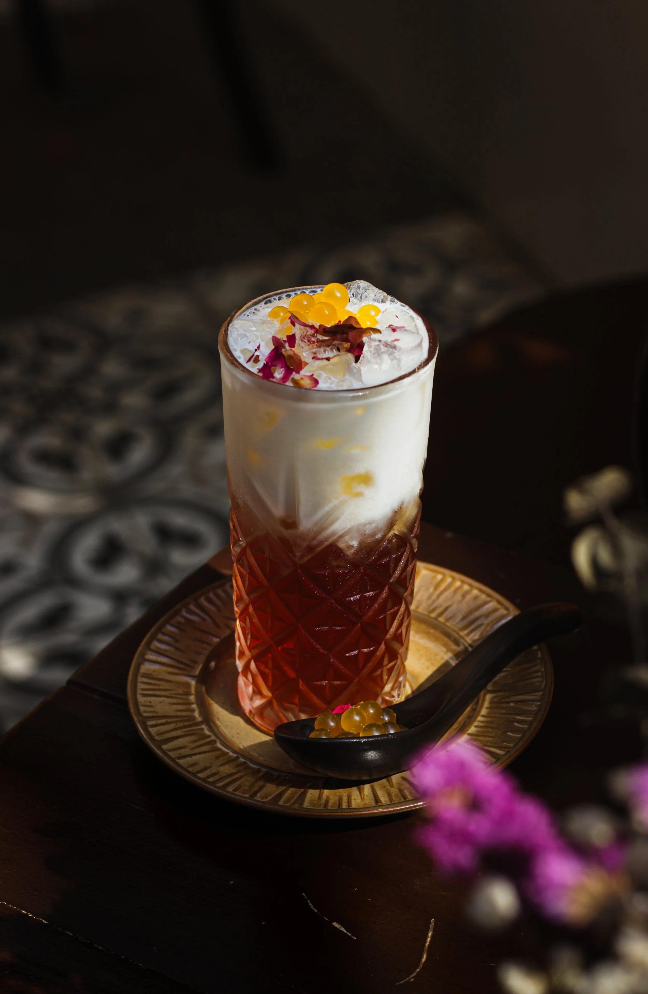 A tall glass of layered colorful beverage garnished with yellow and pink edible flowers, served on a decorative golden tray with a black spoon and yellow berries, set on a dark wooden surface.