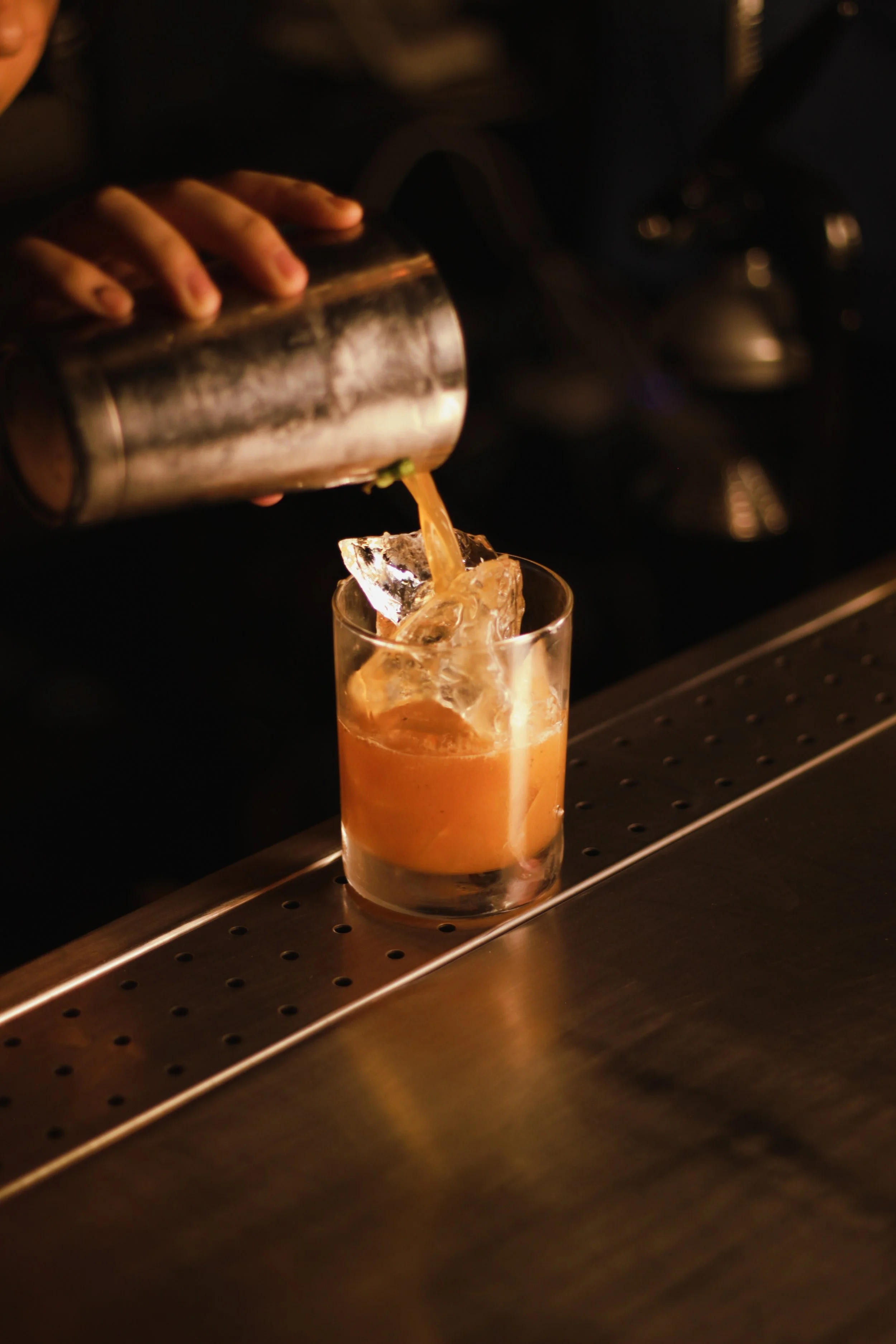 A bartender pours a drink over ice in a glass, with a metal shaker in hand. The setting appears to be a bar with a dark background and metallic surface.