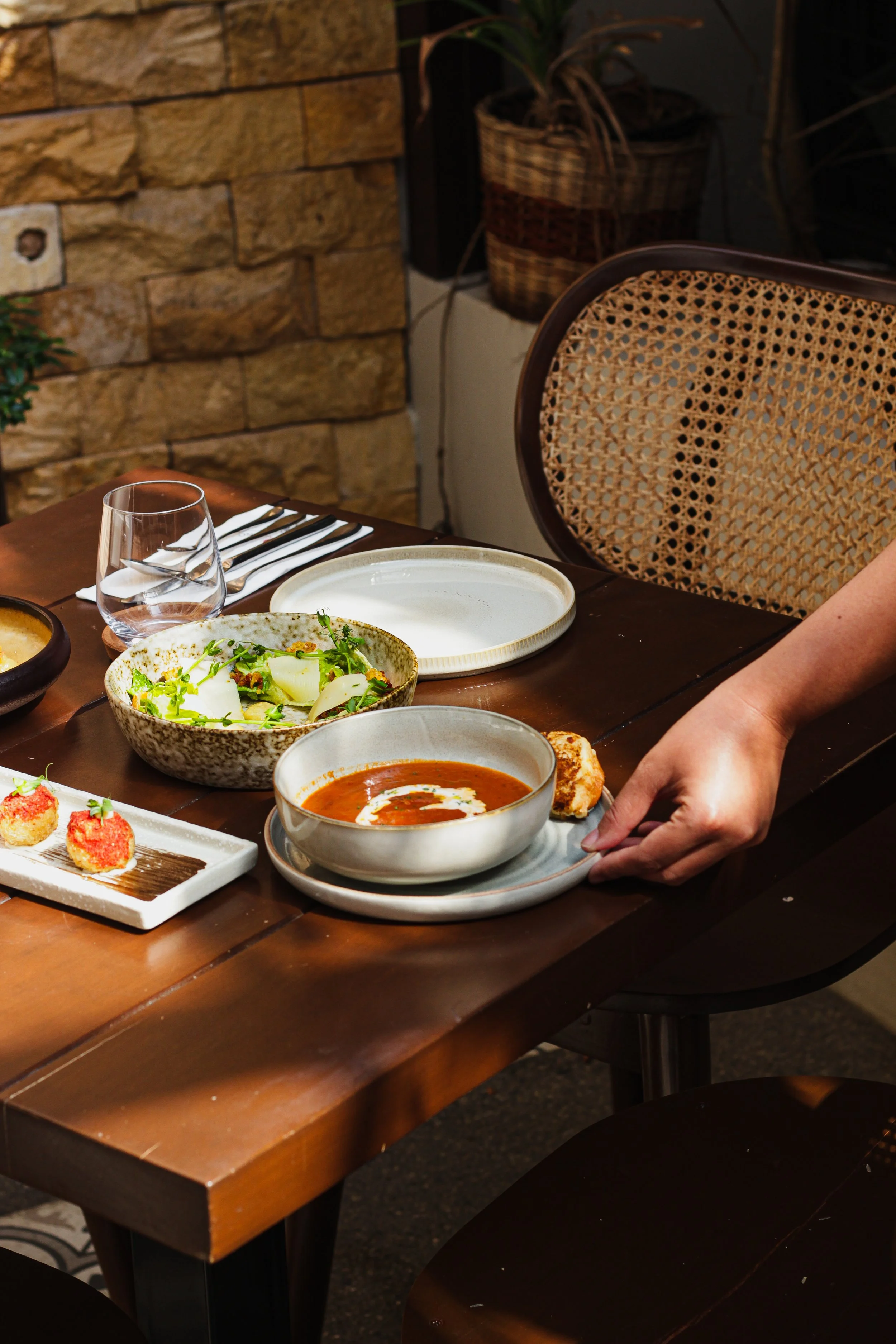 A dining table with various dishes including soup, salad, and appetizers, set with utensils and glassware at a restaurant with a brick wall and woven-style chair in the background.