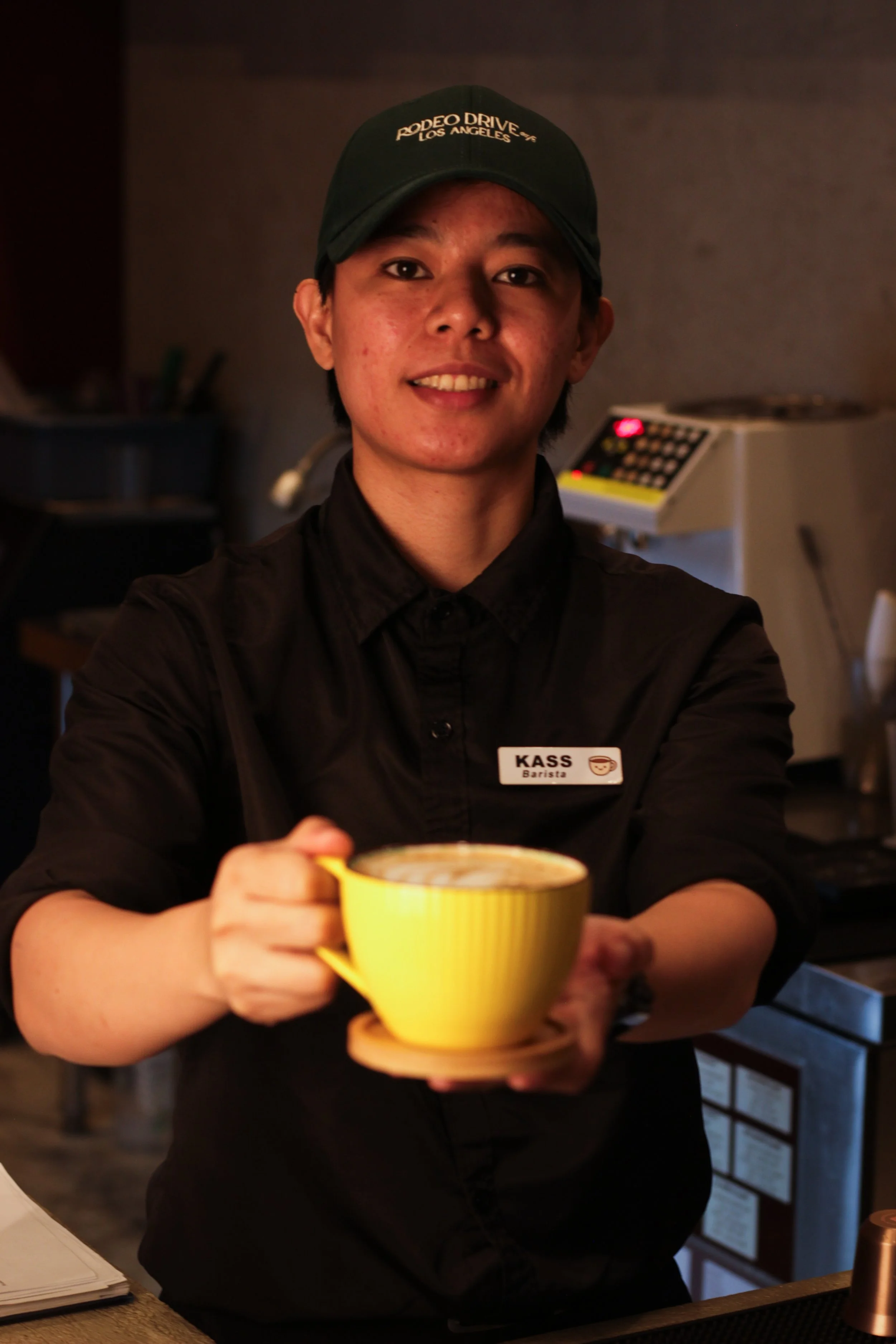A smiling barista wearing a black uniform and cap with 'Rodeo Drive Los Angeles' embroidered on it, holding a yellow mug with a beverage, standing behind a coffee counter in a cafe.